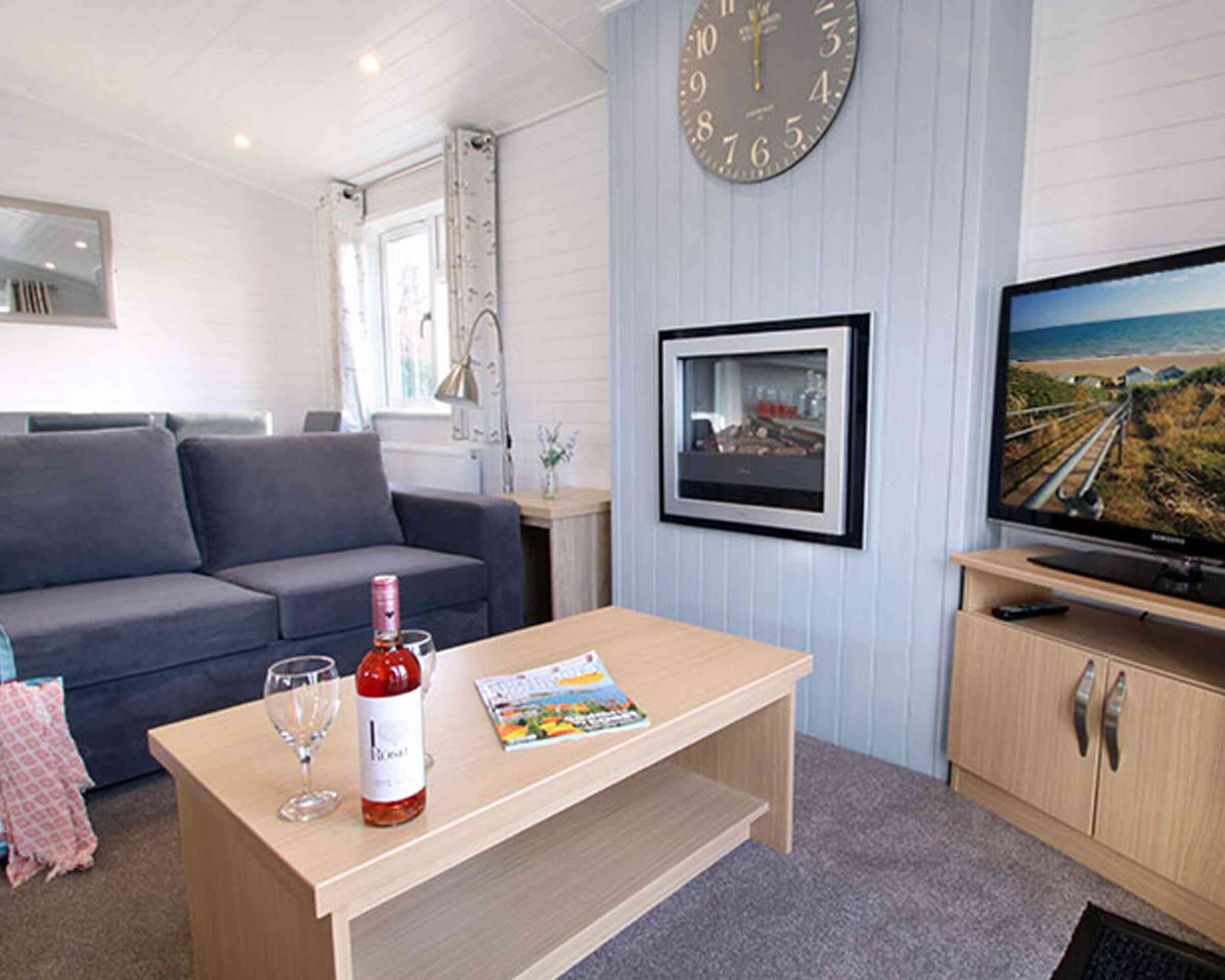 A cozy living room featuring a grey sofa, a coffee table with a bottle of rosé wine and two wine glasses, and a television displaying a scenic view. A clock hangs on the wall, and there are large windows allowing natural light.