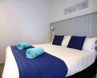 A neatly made bed with white linens and a blue bedspread, featuring two blue towels rolled on top. The room has light-colored walls and a decorative framed artwork above the bed.