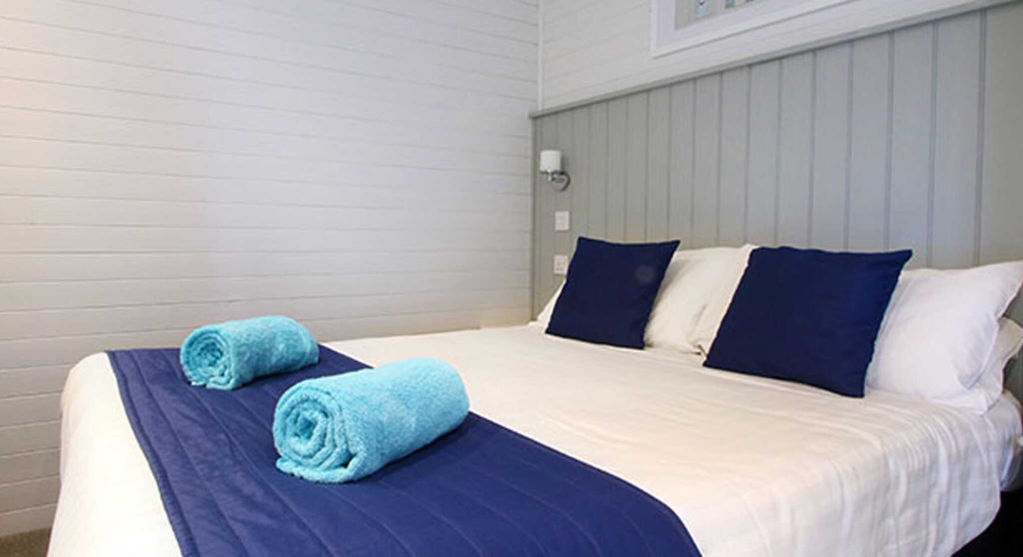 A neatly made bed with white linens and a blue bedspread, featuring two blue towels rolled on top. The room has light-colored walls and a decorative framed artwork above the bed.