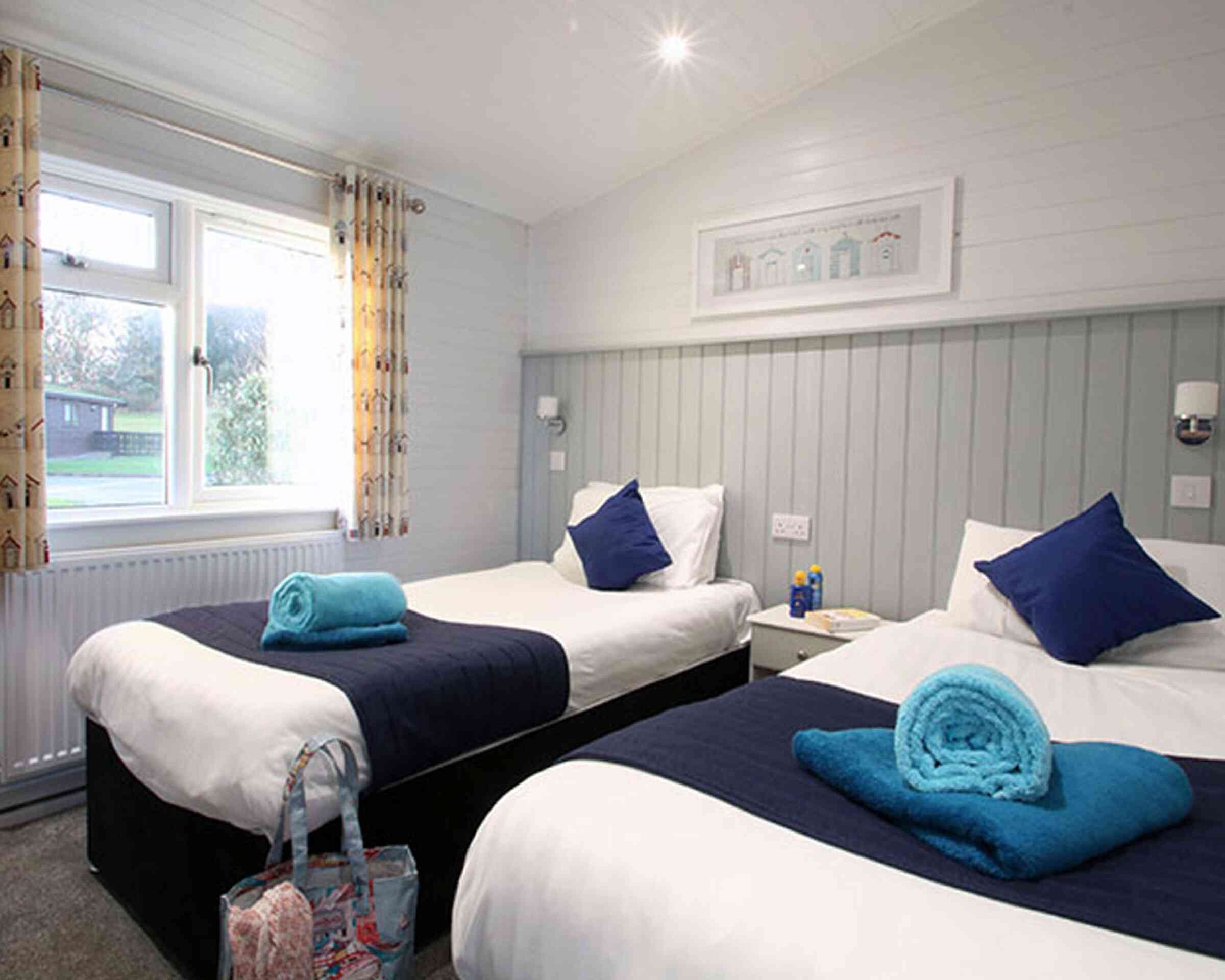 A cozy hotel room featuring two twin beds with navy blue and white bedding. Each bed has a neatly rolled turquoise towel and a decorative blue pillow. Light curtains frame a window that lets in natural light, illuminating the room's light gray walls and soft carpet. A small nightstand and a stylish bag are placed by one bed.