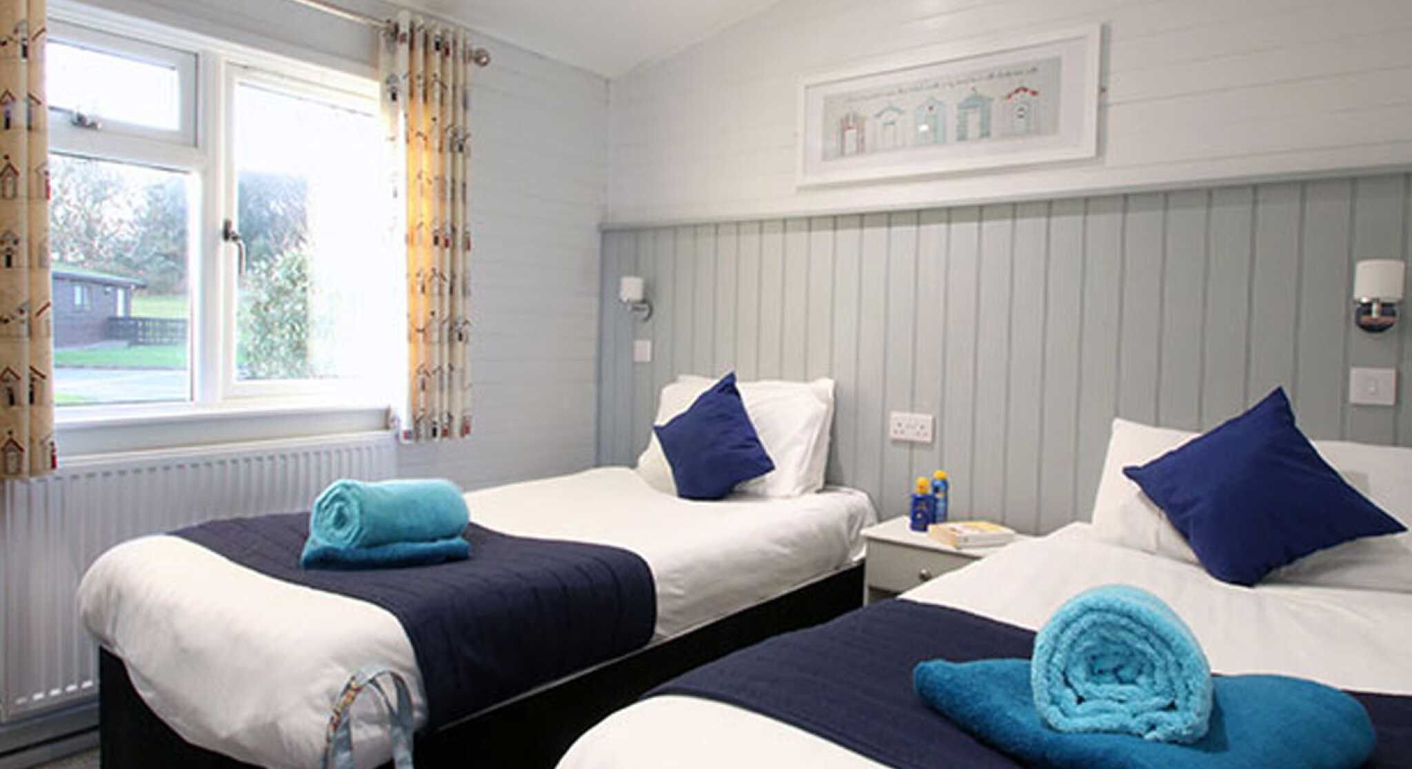 A cozy hotel room featuring two twin beds with navy blue and white bedding. Each bed has a neatly rolled turquoise towel and a decorative blue pillow. Light curtains frame a window that lets in natural light, illuminating the room's light gray walls and soft carpet. A small nightstand and a stylish bag are placed by one bed.