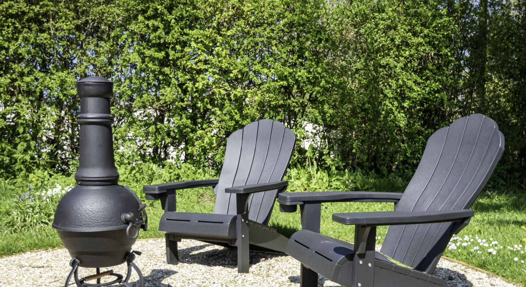 Two black Adirondack chairs are positioned beside a cast-iron chiminea, surrounded by a gravel area and lush green hedges in the background. The scene is bright, suggesting a clear, sunny day.