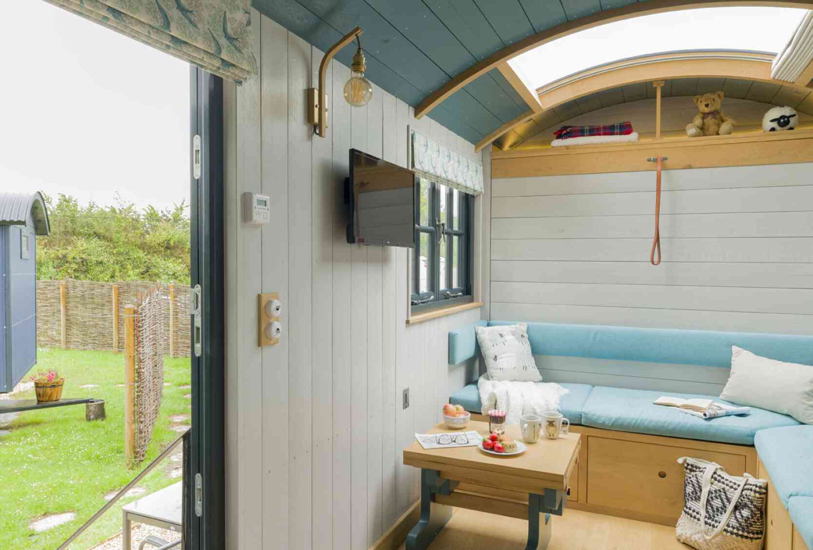 Cozy interior of a tiny house featuring a light blue sofa, a wooden table with snacks, and a mounted television. Large windows provide a view of greenery outside. A teddy bear sits on a shelf, and there are decorative touches on the walls.