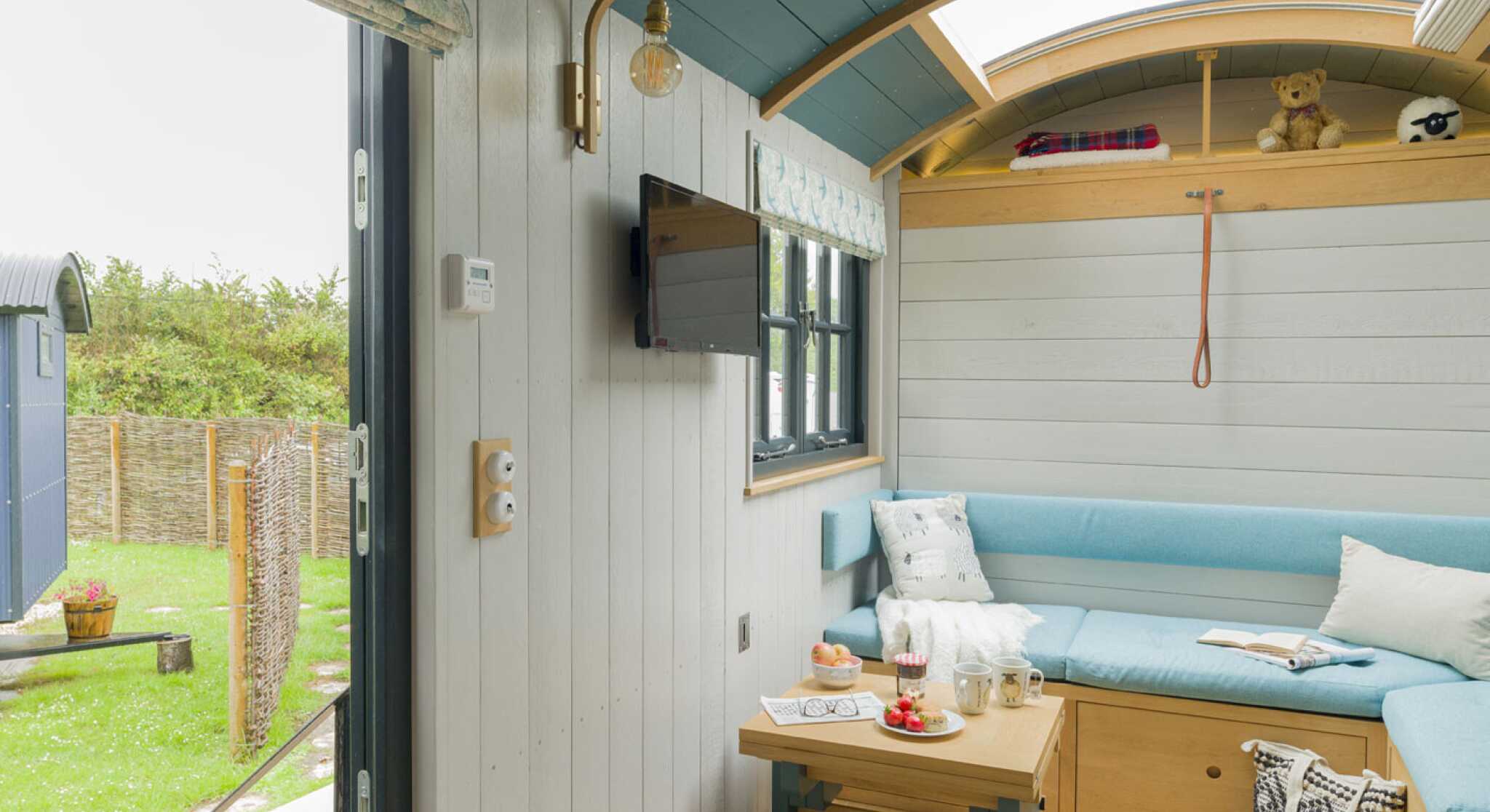 Cozy interior of a tiny house featuring a light blue sofa, a wooden table with snacks, and a mounted television. Large windows provide a view of greenery outside. A teddy bear sits on a shelf, and there are decorative touches on the walls.