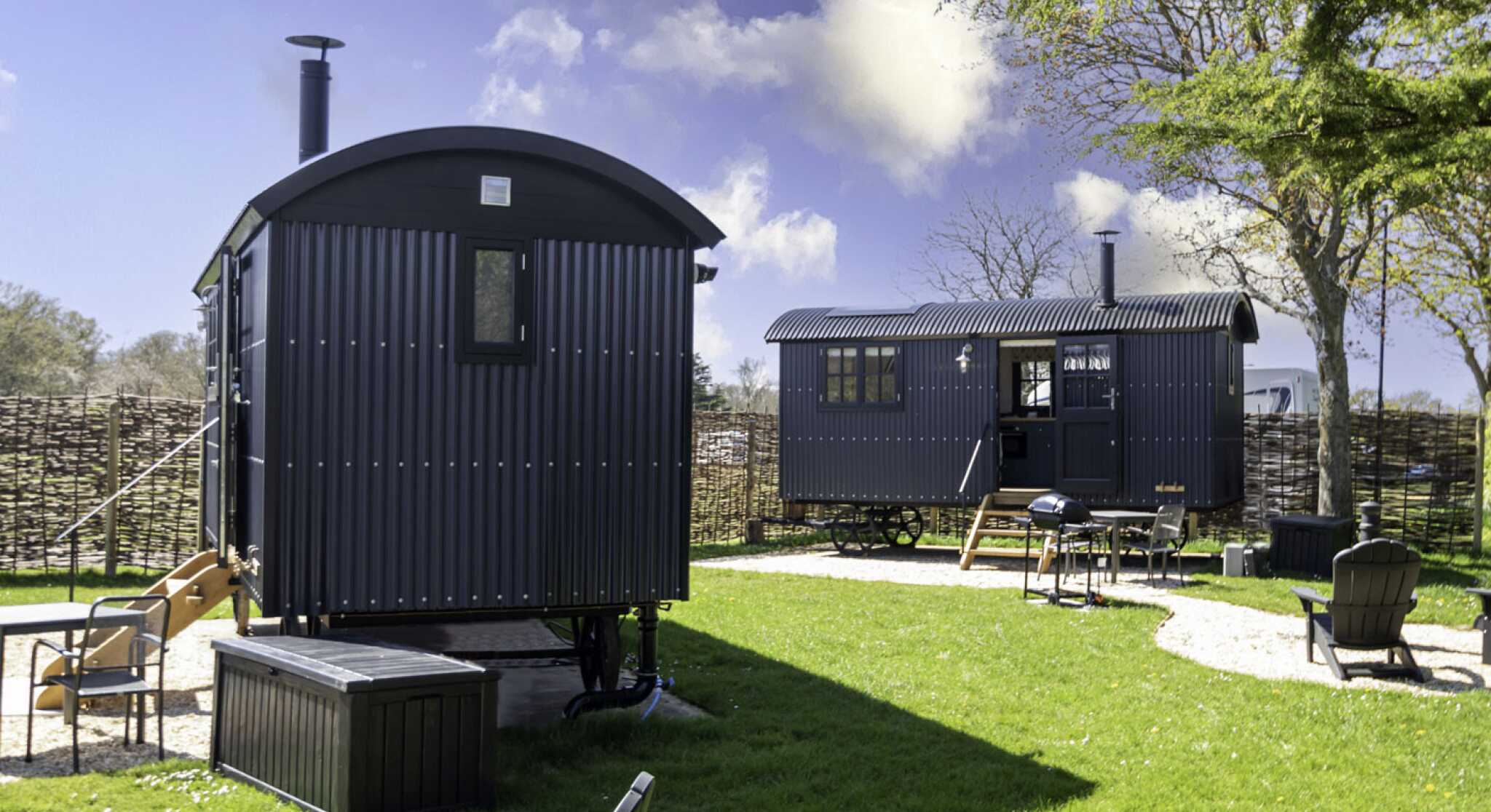 Two dark-colored shepherd's huts sit in a grassy area. One hut is slightly larger with a porch, while the other is smaller. There's a fire pit and several outdoor chairs nearby, surrounded by a rustic wooden fence and trees under a partly cloudy sky.