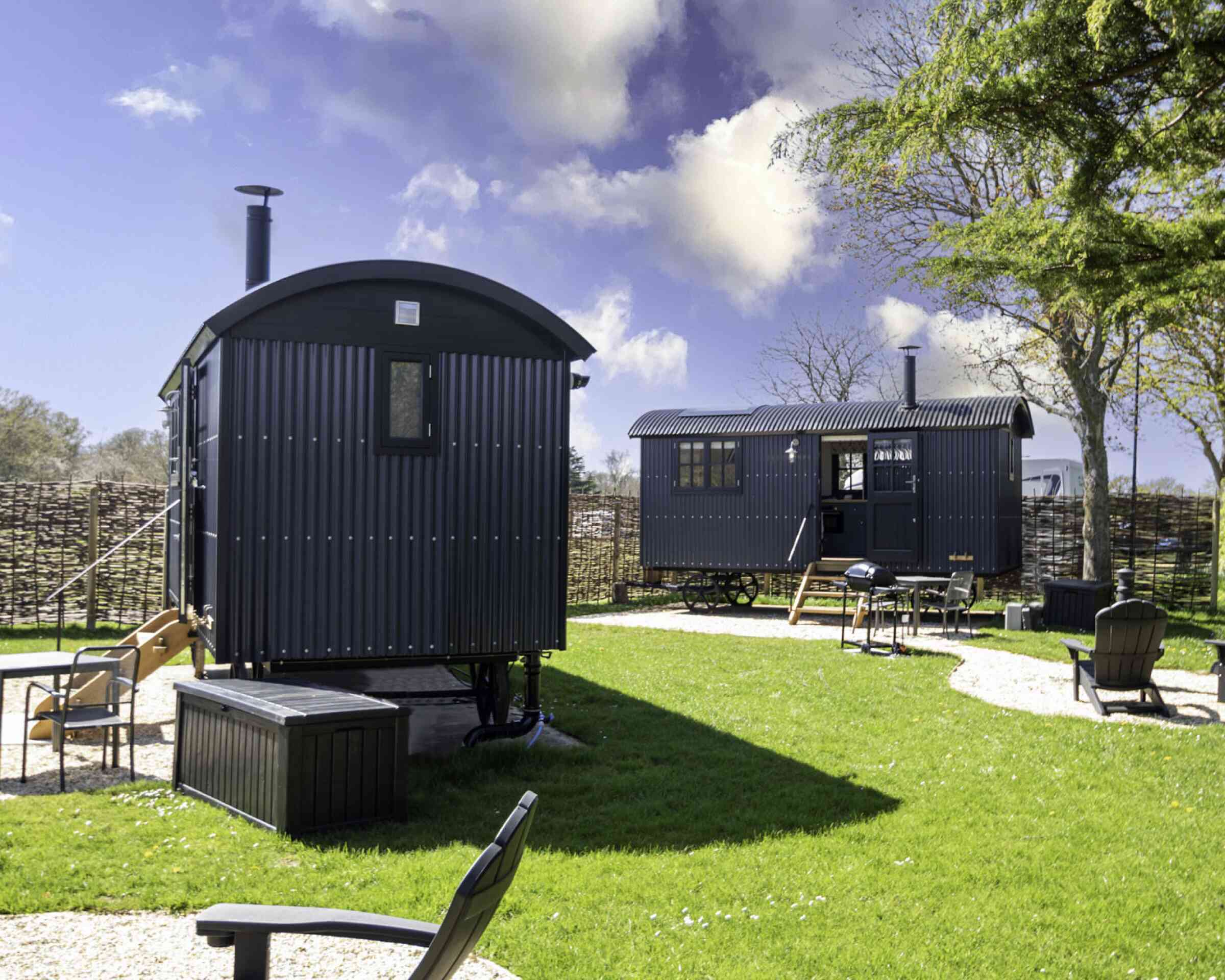Two dark-colored shepherd's huts sit in a grassy area. One hut is slightly larger with a porch, while the other is smaller. There's a fire pit and several outdoor chairs nearby, surrounded by a rustic wooden fence and trees under a partly cloudy sky.