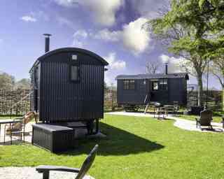 Two dark-colored shepherd's huts sit in a grassy area. One hut is slightly larger with a porch, while the other is smaller. There's a fire pit and several outdoor chairs nearby, surrounded by a rustic wooden fence and trees under a partly cloudy sky.