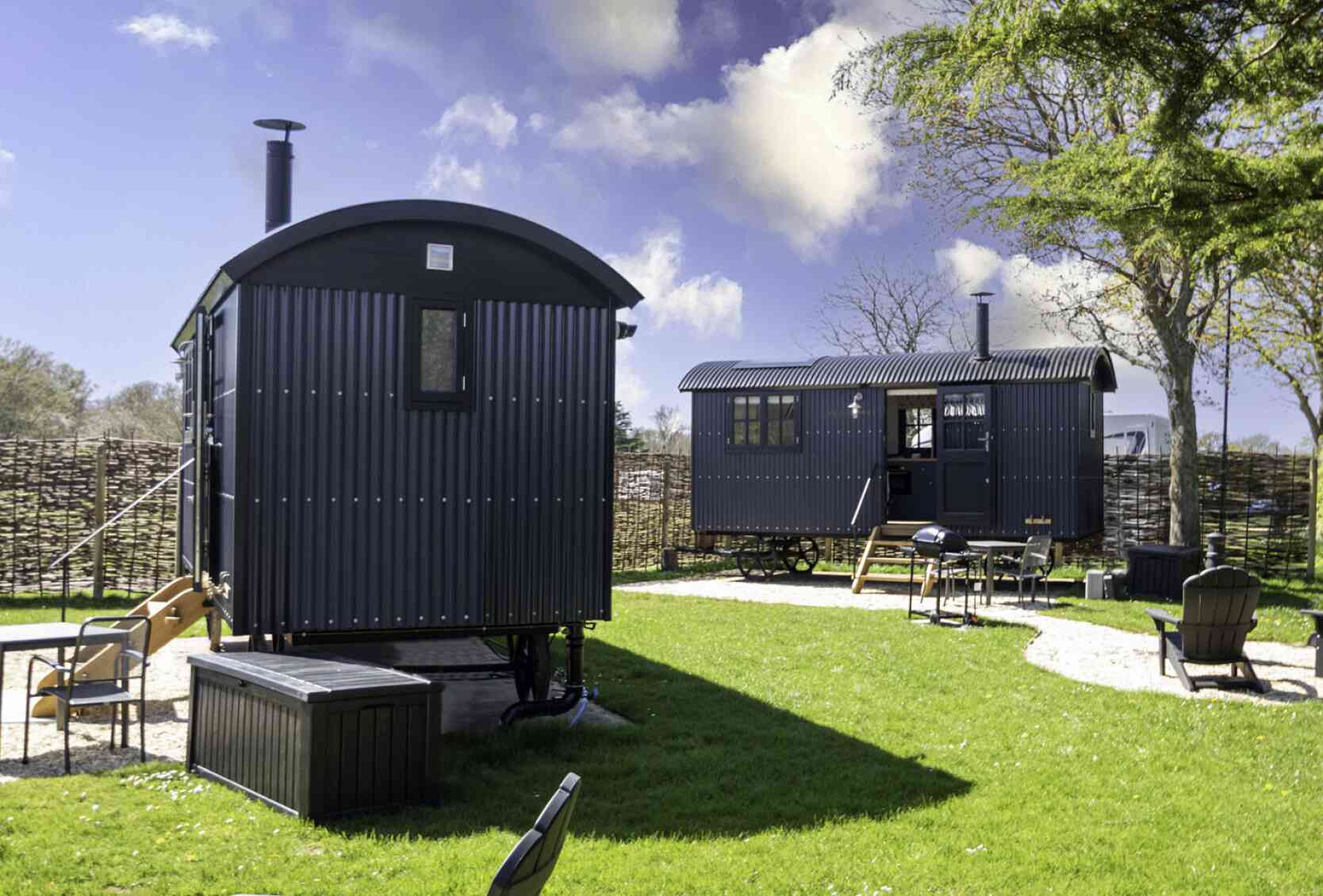 Two dark-colored shepherd's huts sit in a grassy area. One hut is slightly larger with a porch, while the other is smaller. There's a fire pit and several outdoor chairs nearby, surrounded by a rustic wooden fence and trees under a partly cloudy sky.