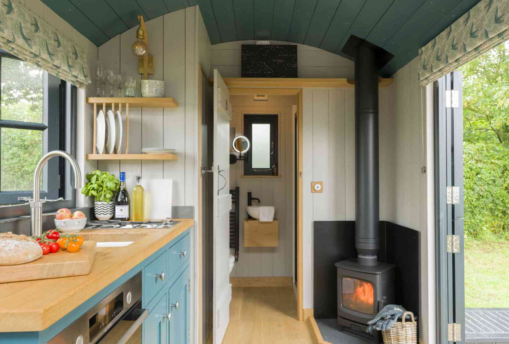 A cozy tiny home interior featuring a small kitchen with teal cabinets, a wooden countertop, and stainless steel appliances. A wood-burning stove is visible in the corner, with a warm glow. Natural light enters through a window, highlighting the simple yet functional design. Greenery can be seen outside through the door.