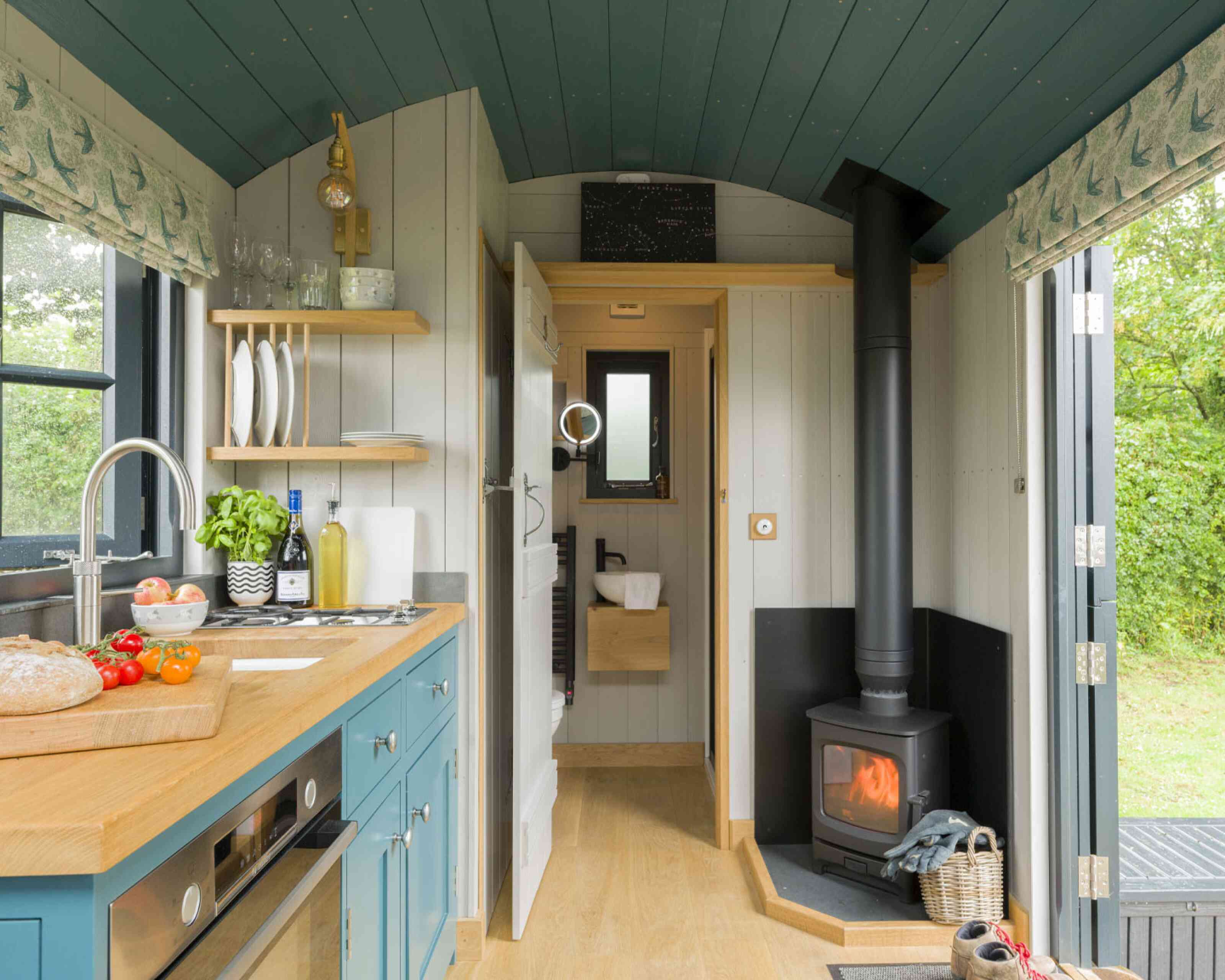 A cozy tiny home interior featuring a small kitchen with teal cabinets, a wooden countertop, and stainless steel appliances. A wood-burning stove is visible in the corner, with a warm glow. Natural light enters through a window, highlighting the simple yet functional design. Greenery can be seen outside through the door.