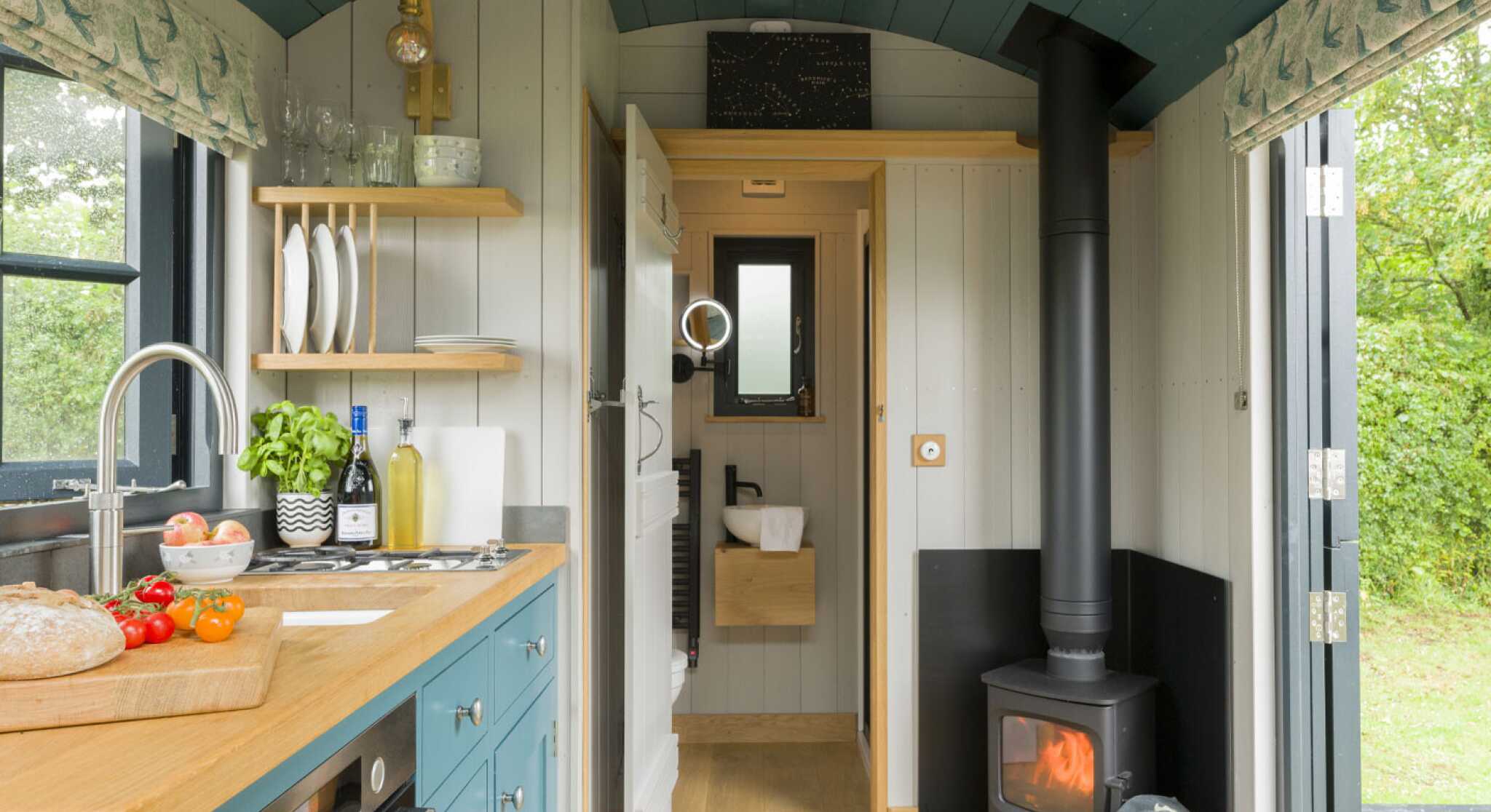 A cozy tiny home interior featuring a small kitchen with teal cabinets, a wooden countertop, and stainless steel appliances. A wood-burning stove is visible in the corner, with a warm glow. Natural light enters through a window, highlighting the simple yet functional design. Greenery can be seen outside through the door.
