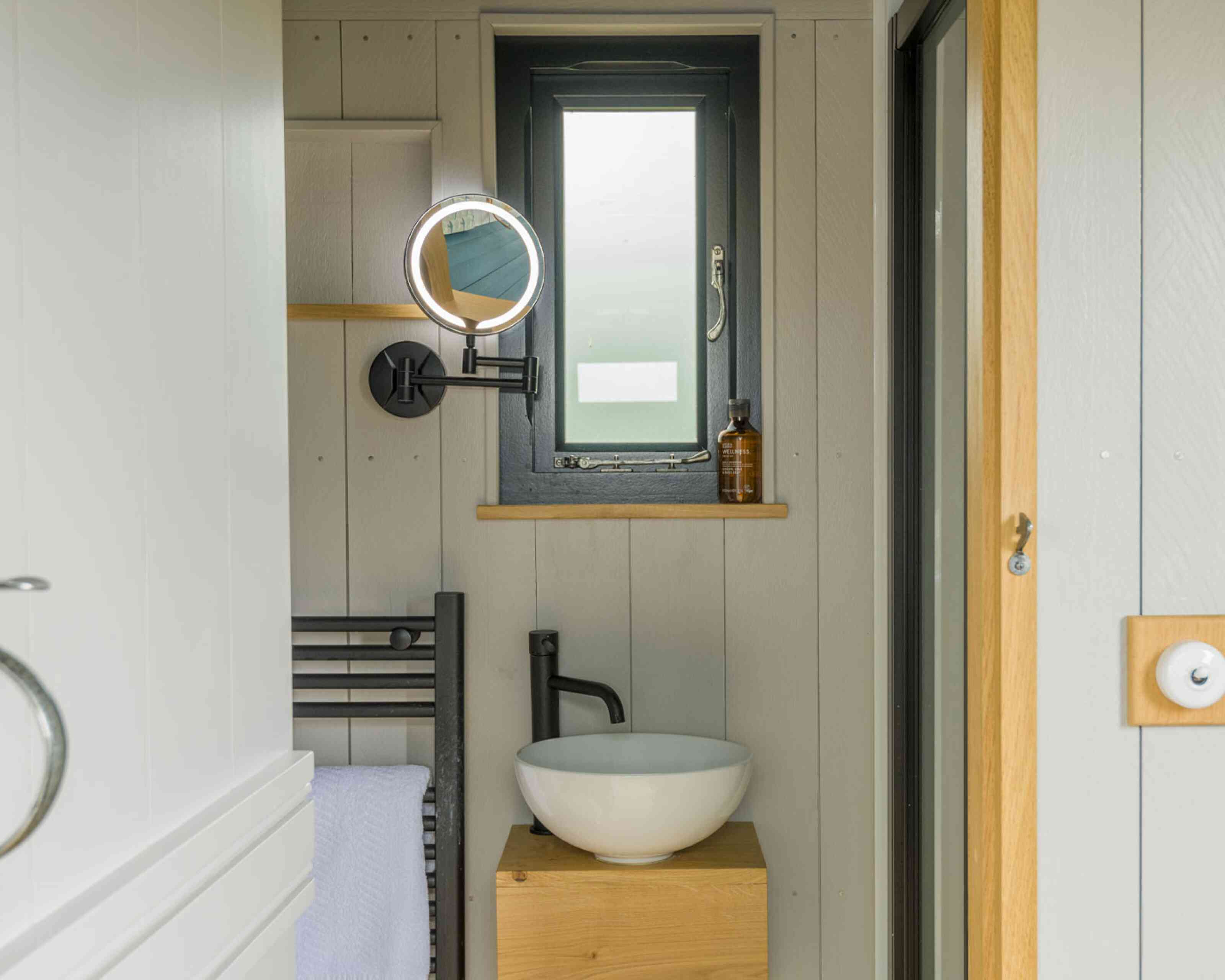 A minimalist bathroom featuring a small round window, a mirror with built-in lighting, a modern basin atop a wooden stand, a black towel rack, and a shelf with a bottle. The walls are light-colored wood, creating a clean and modern aesthetic.