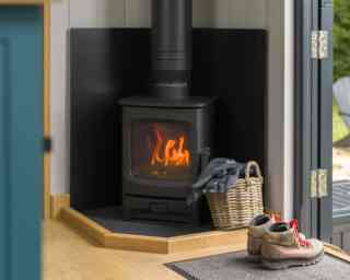 A black wood stove emits a warm glow, positioned in a cozy corner with a wooden floor. A woven basket filled with a blanket sits nearby, alongside a pair of rugged boots on a mat. Natural light filters in from an open door, adding to the inviting atmosphere.