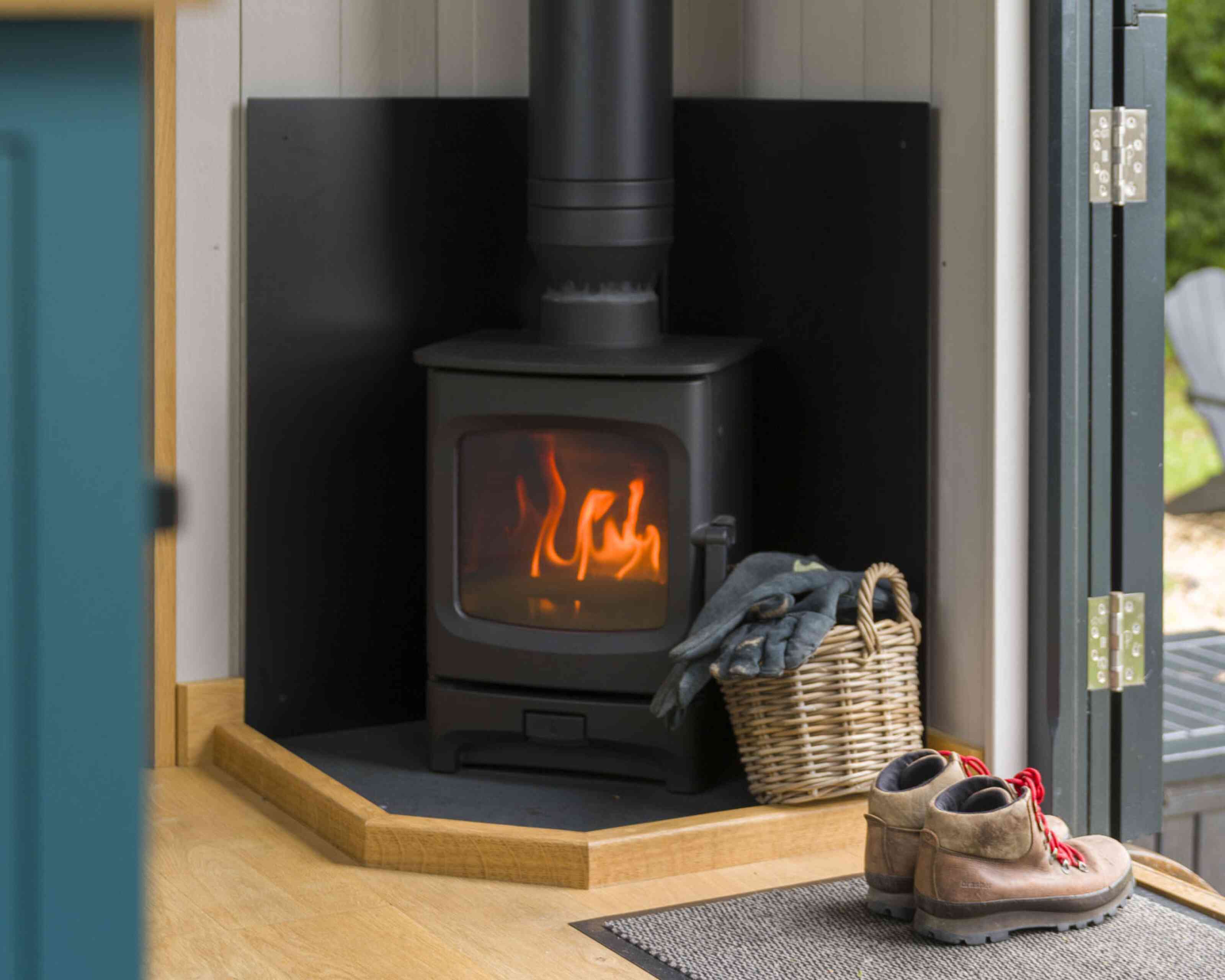 A black wood stove emits a warm glow, positioned in a cozy corner with a wooden floor. A woven basket filled with a blanket sits nearby, alongside a pair of rugged boots on a mat. Natural light filters in from an open door, adding to the inviting atmosphere.