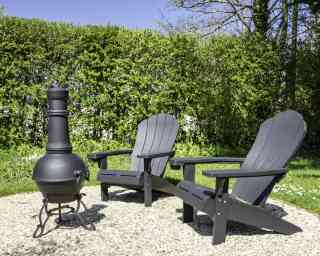 Two black wooden Adirondack chairs are positioned around a chiminea in a garden setting. The chiminea is made of metal and sits on a stone surface. Lush green hedges and plants surround the area under a clear blue sky.