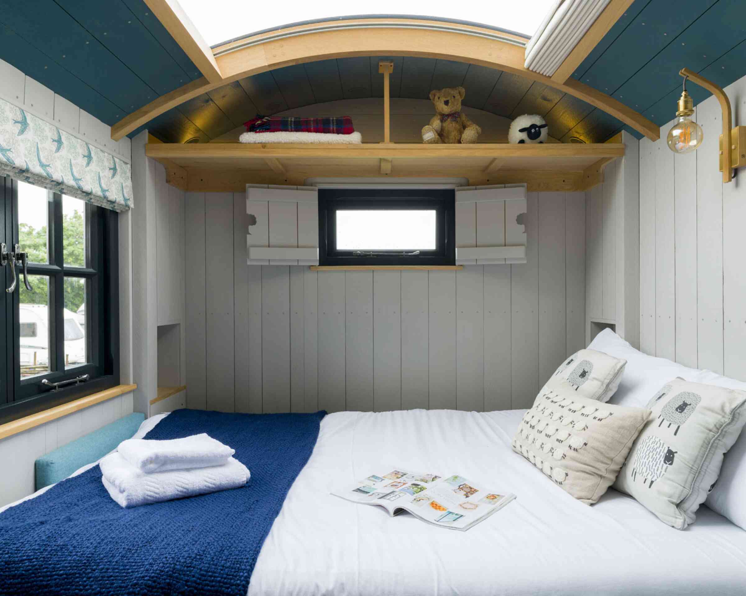 Cozy interior of a small bedroom featuring a double bed with white linens, a blue crocheted blanket, and decorative pillows. A teddy bear and a sheep figurine are on a wooden shelf above the bed. A small window with a white frame provides natural light, and a stylish wall lamp adds warmth to the space.