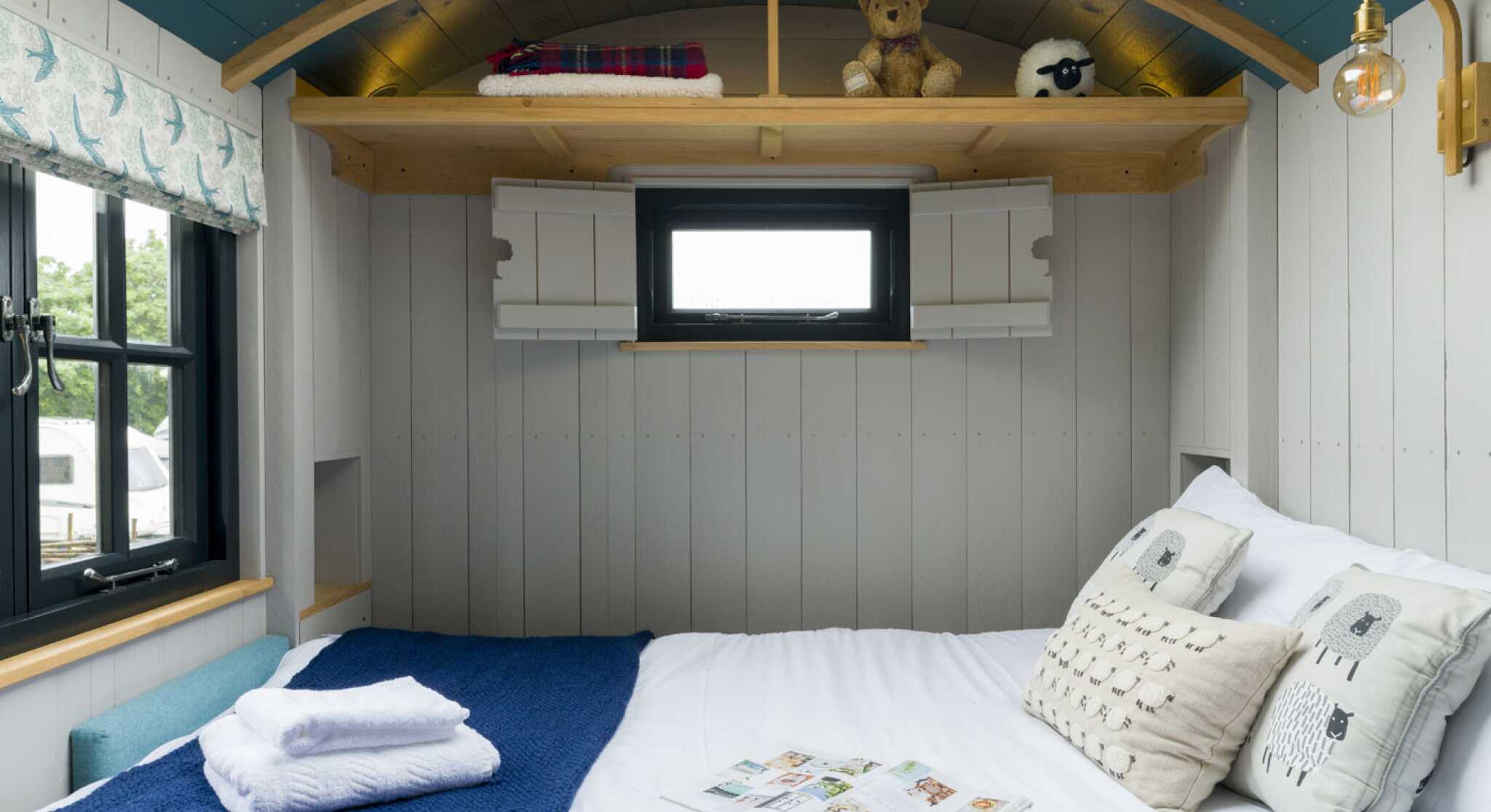 Cozy interior of a small bedroom featuring a double bed with white linens, a blue crocheted blanket, and decorative pillows. A teddy bear and a sheep figurine are on a wooden shelf above the bed. A small window with a white frame provides natural light, and a stylish wall lamp adds warmth to the space.