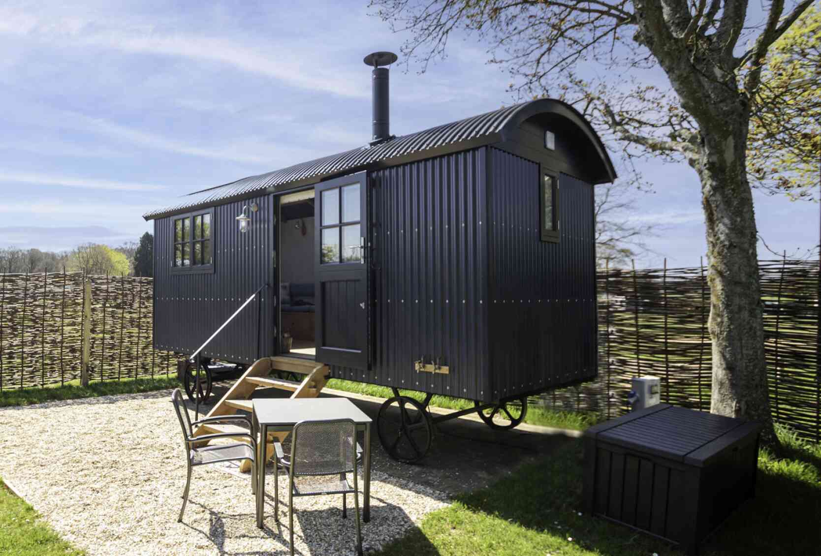 A black, rustic caravan with a rounded roof is parked on a grassy area surrounded by a woven fence. In front, there’s a small wooden table with chairs, and a storage box sits nearby. The background features a clear blue sky and trees.