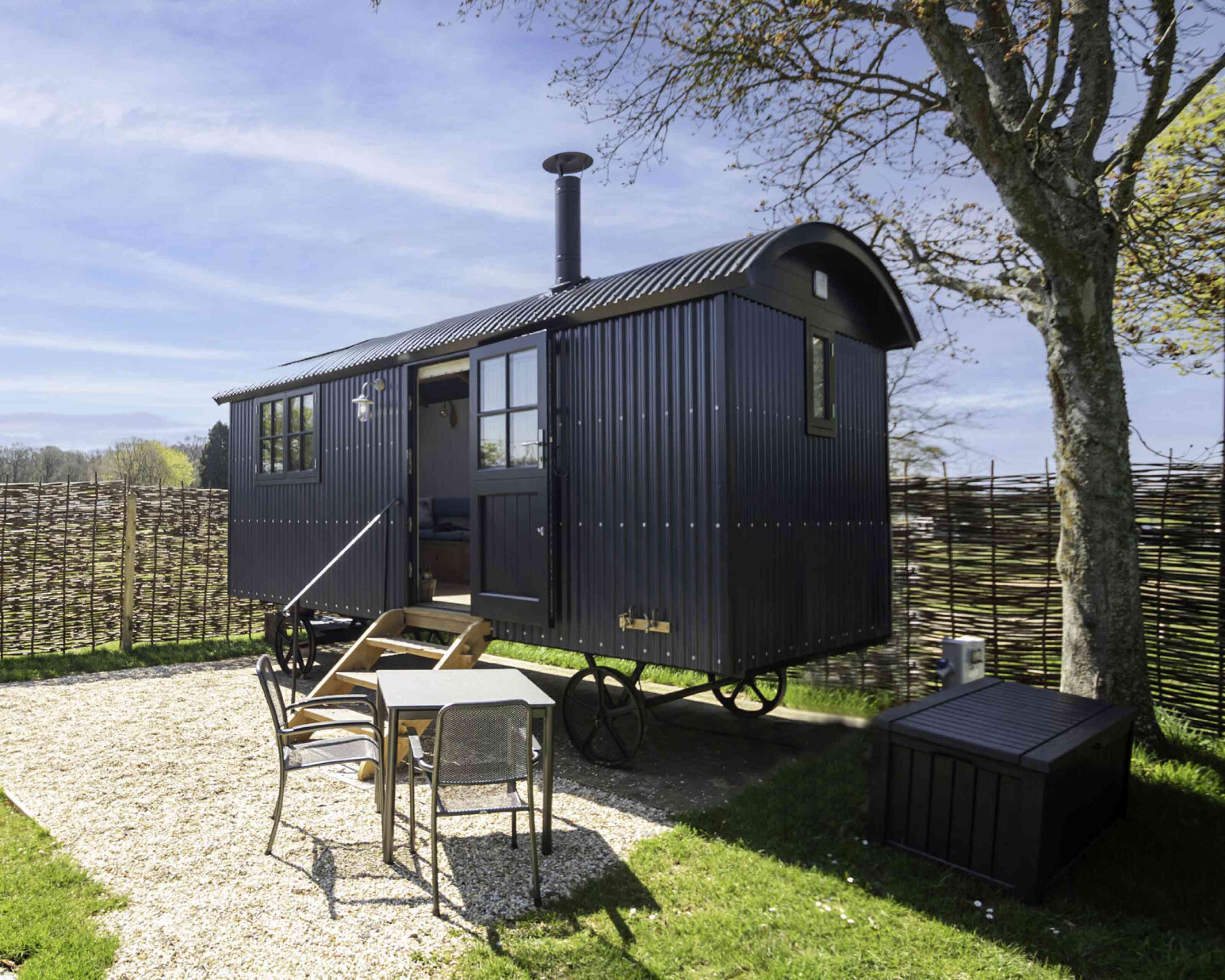 A black, rustic caravan with a rounded roof is parked on a grassy area surrounded by a woven fence. In front, there’s a small wooden table with chairs, and a storage box sits nearby. The background features a clear blue sky and trees.