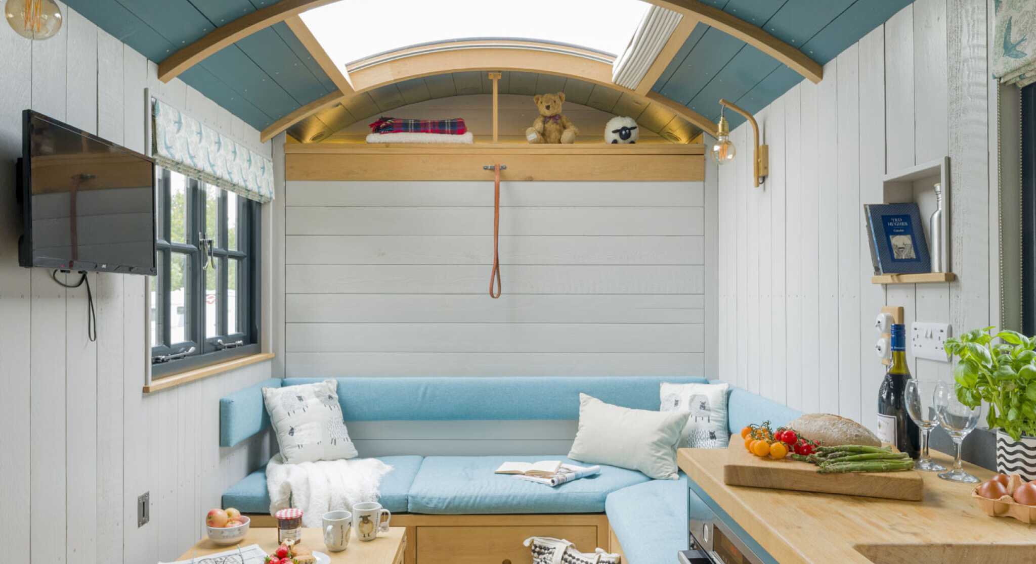 A cozy interior of a tiny home featuring a light blue sectional sofa, a wooden coffee table with snacks and drinks, and an open kitchen area. Natural light comes through a skylight, highlighting the wooden ceiling and white walls. Decorative elements include a teddy bear and houseplants. A television is mounted on the wall.