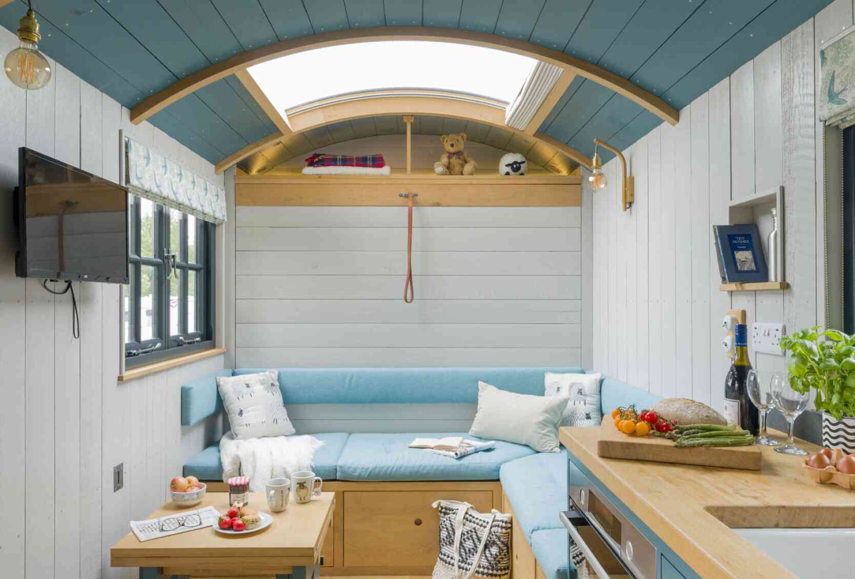 A cozy interior of a tiny home featuring a light blue sectional sofa, a wooden coffee table with snacks and drinks, and an open kitchen area. Natural light comes through a skylight, highlighting the wooden ceiling and white walls. Decorative elements include a teddy bear and houseplants. A television is mounted on the wall.