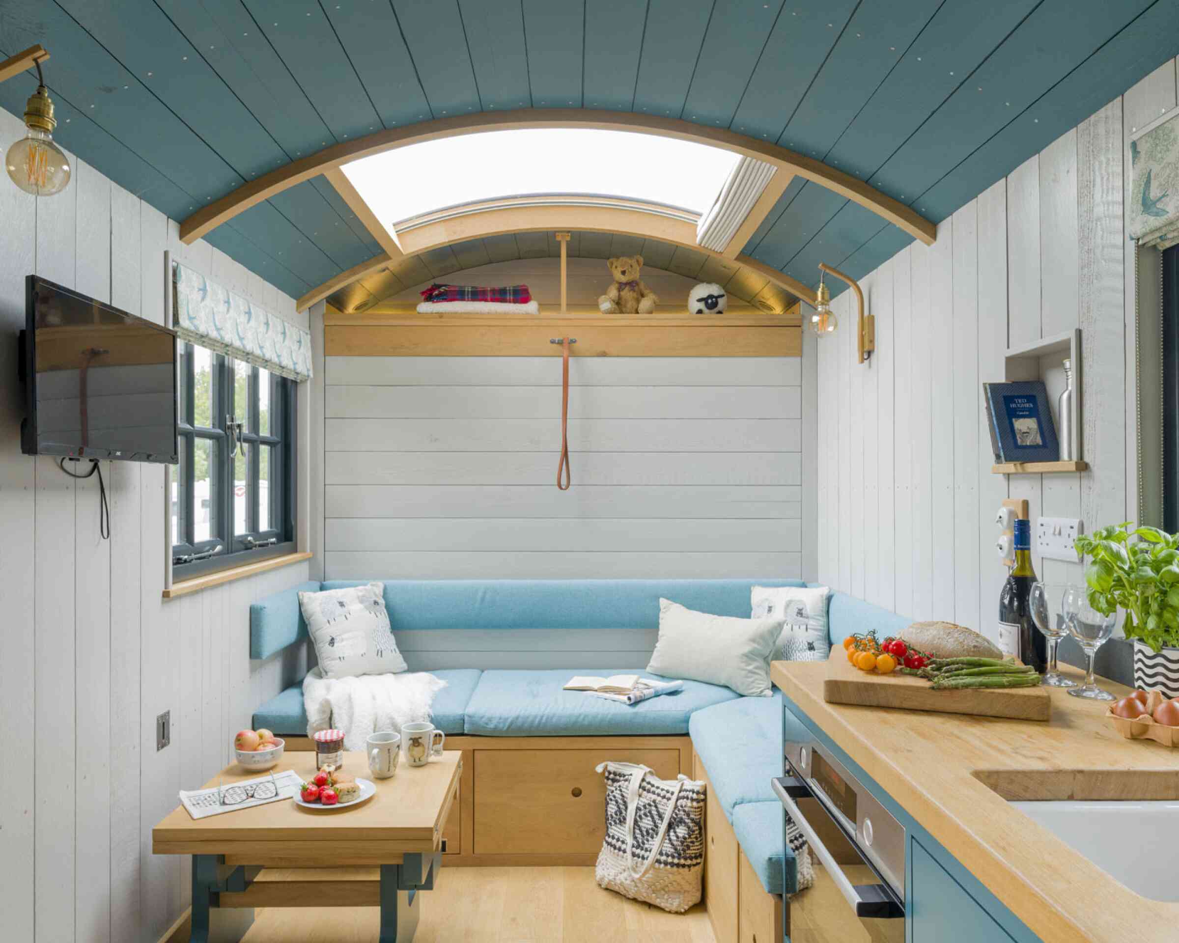 A cozy interior of a tiny home featuring a light blue sectional sofa, a wooden coffee table with snacks and drinks, and an open kitchen area. Natural light comes through a skylight, highlighting the wooden ceiling and white walls. Decorative elements include a teddy bear and houseplants. A television is mounted on the wall.