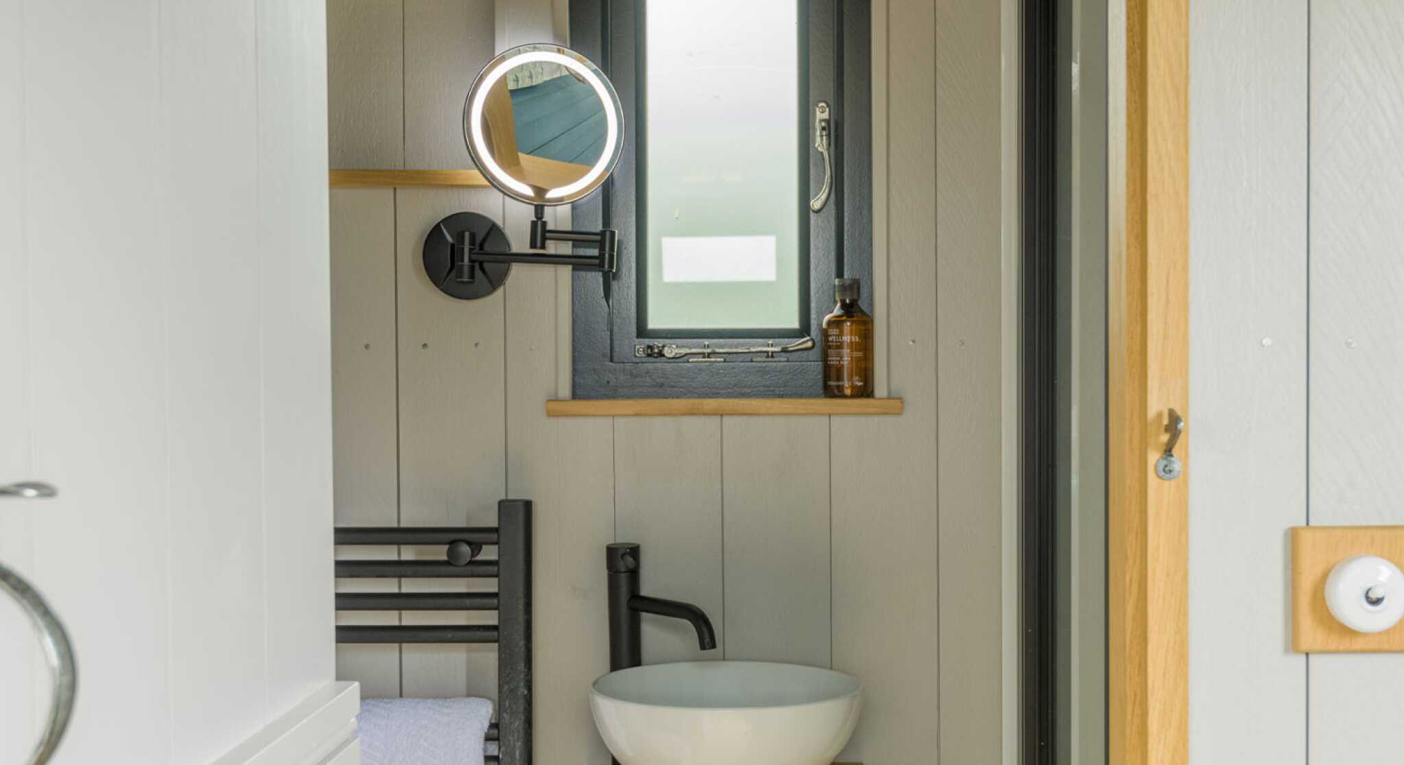 Compact bathroom featuring a round mirror, a small window, a stylish sink atop a wooden stand, and a towel rack. The walls are paneled in light wood, creating a modern and airy feel.