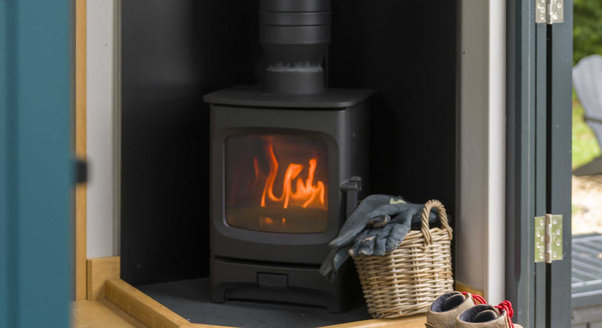 A cozy wood-burning stove emits a warm glow, situated in a corner with a black backing. A wicker basket sits nearby, and a pair of worn boots rests on a welcome mat.