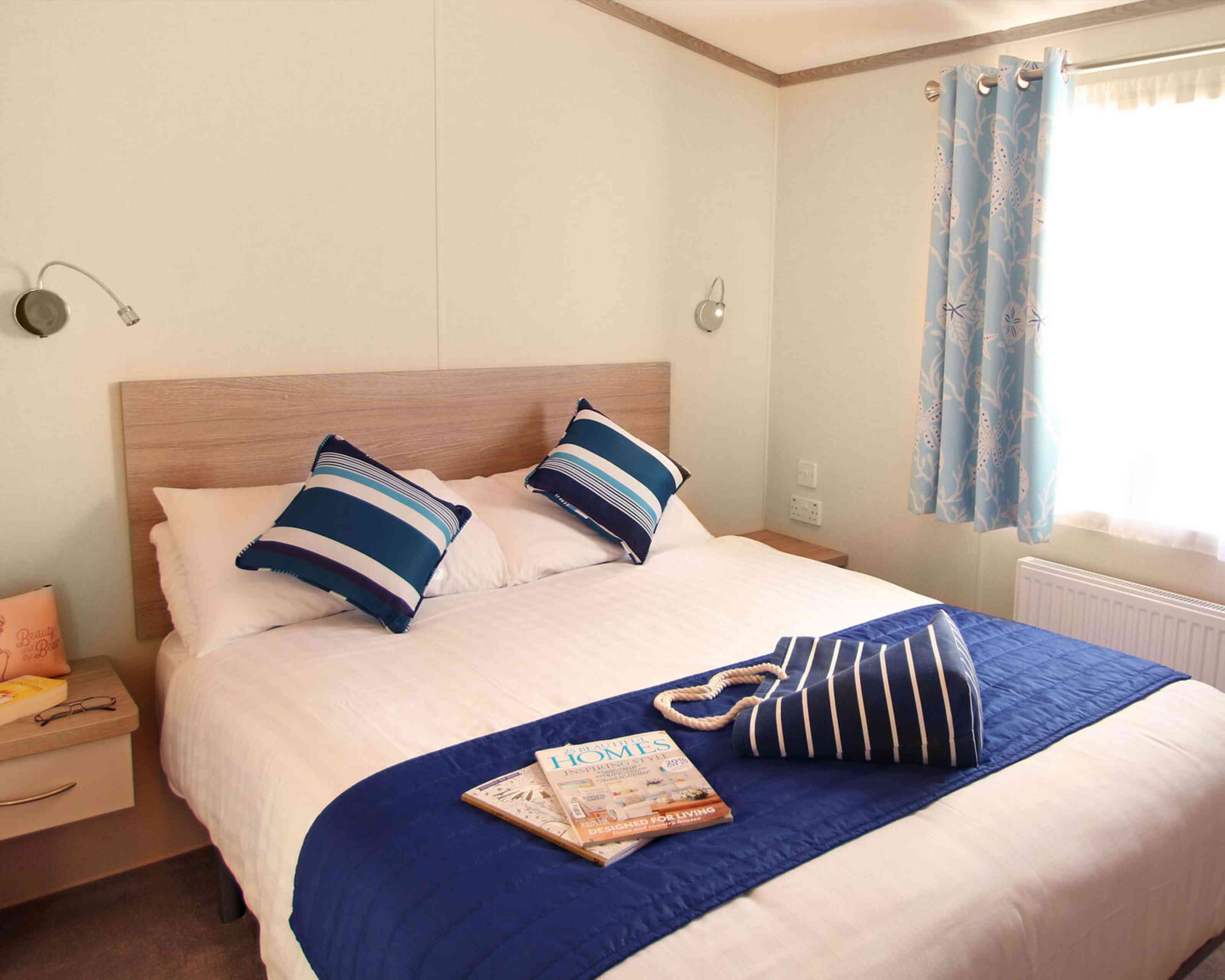 Cozy bedroom featuring a large bed with blue and white striped pillows, a navy blue throw, and a bedside table. Natural light streams through a window adorned with light blue curtains. A magazine and a striped bag are placed on the bed.
