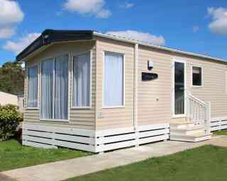 A modern mobile home with large windows, white shutters, and a small porch, surrounded by greenery and blue sky.