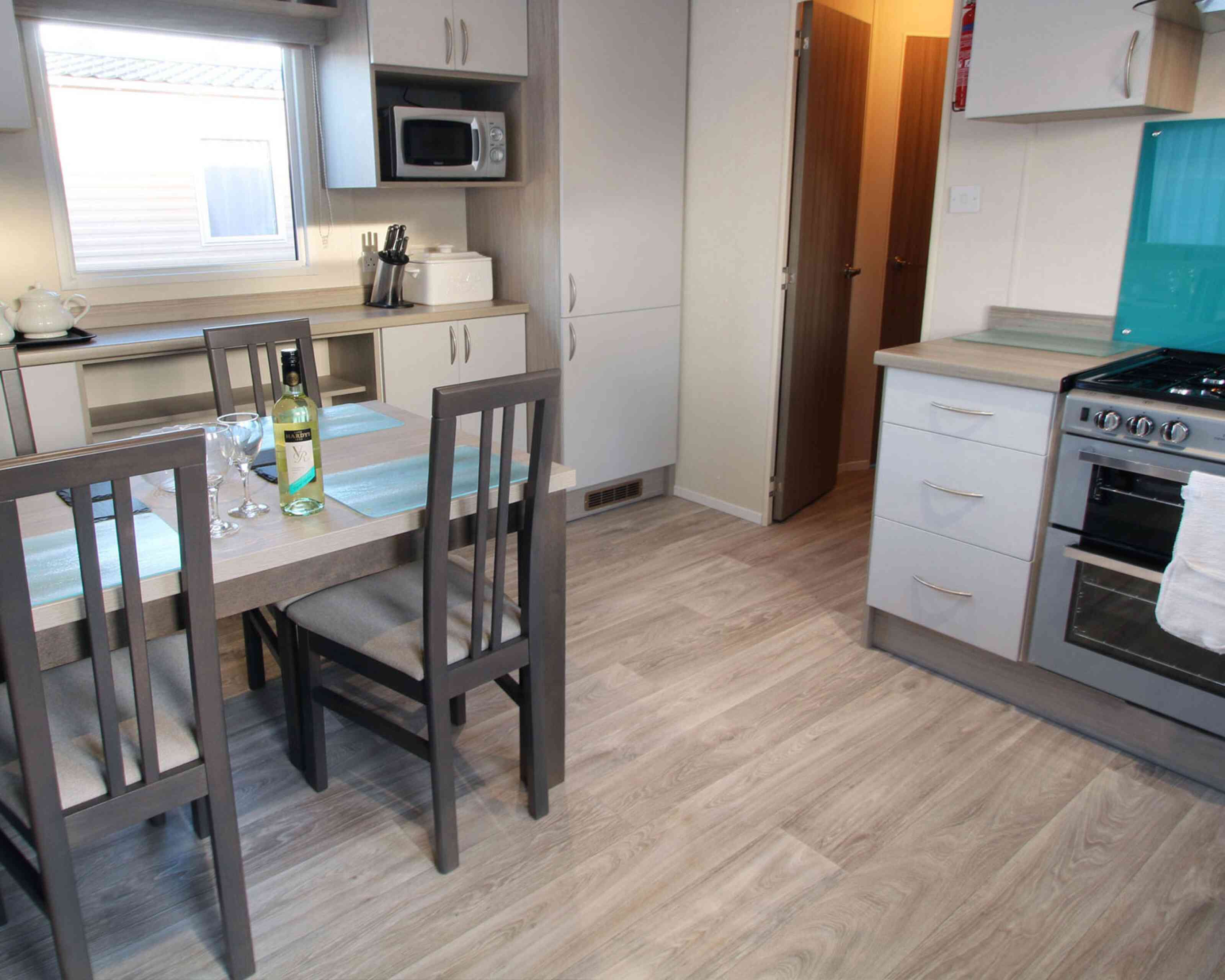 Modern kitchen featuring a dining area with a table set for two, including glasses and a bottle of wine. A gas stove with an oven, a microwave, and light wood flooring complement the sleek cabinetry. A door leads to another room in the background.