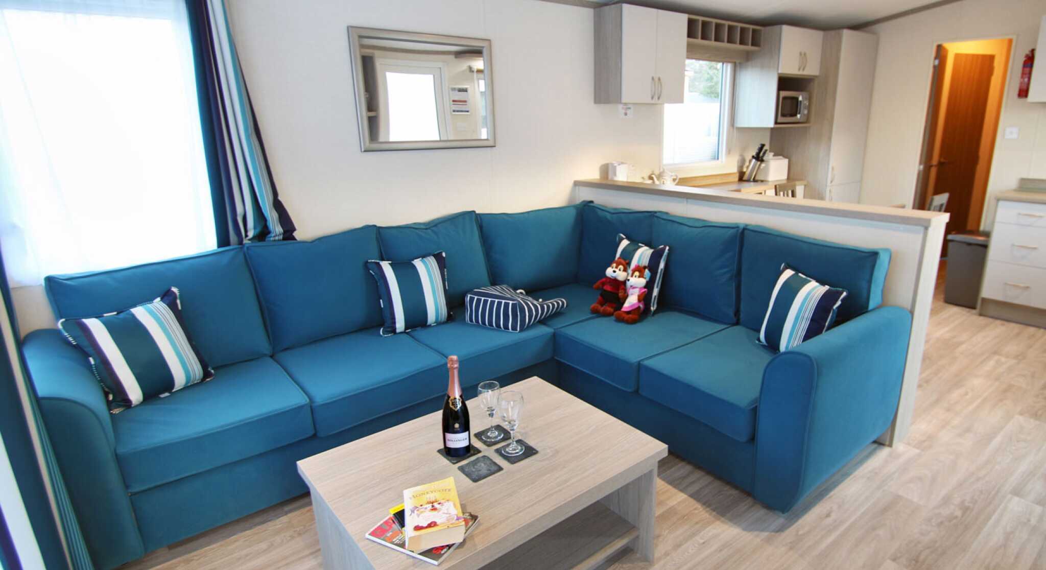 A cozy living area featuring a large blue sectional sofa with striped pillows and a light wooden coffee table. A bottle of champagne and glasses sit on the table, alongside magazines. The space is well-lit with natural light coming through a window, and there are hints of a kitchen in the background.