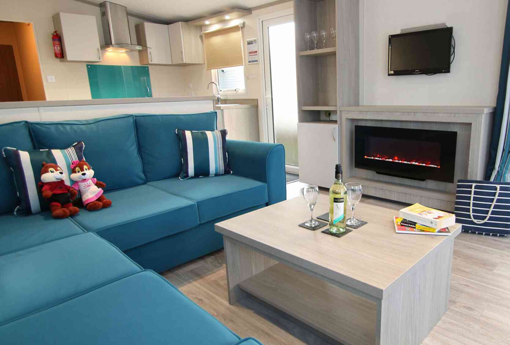 A cozy living room with a teal sectional sofa adorned with blue and white striped cushions. Two plush toys sit on the sofa. A wooden coffee table holds a bottle of wine and two glasses. A modern electric fireplace is below a small wall-mounted TV, with a doorway leading to an outdoor area in the background.