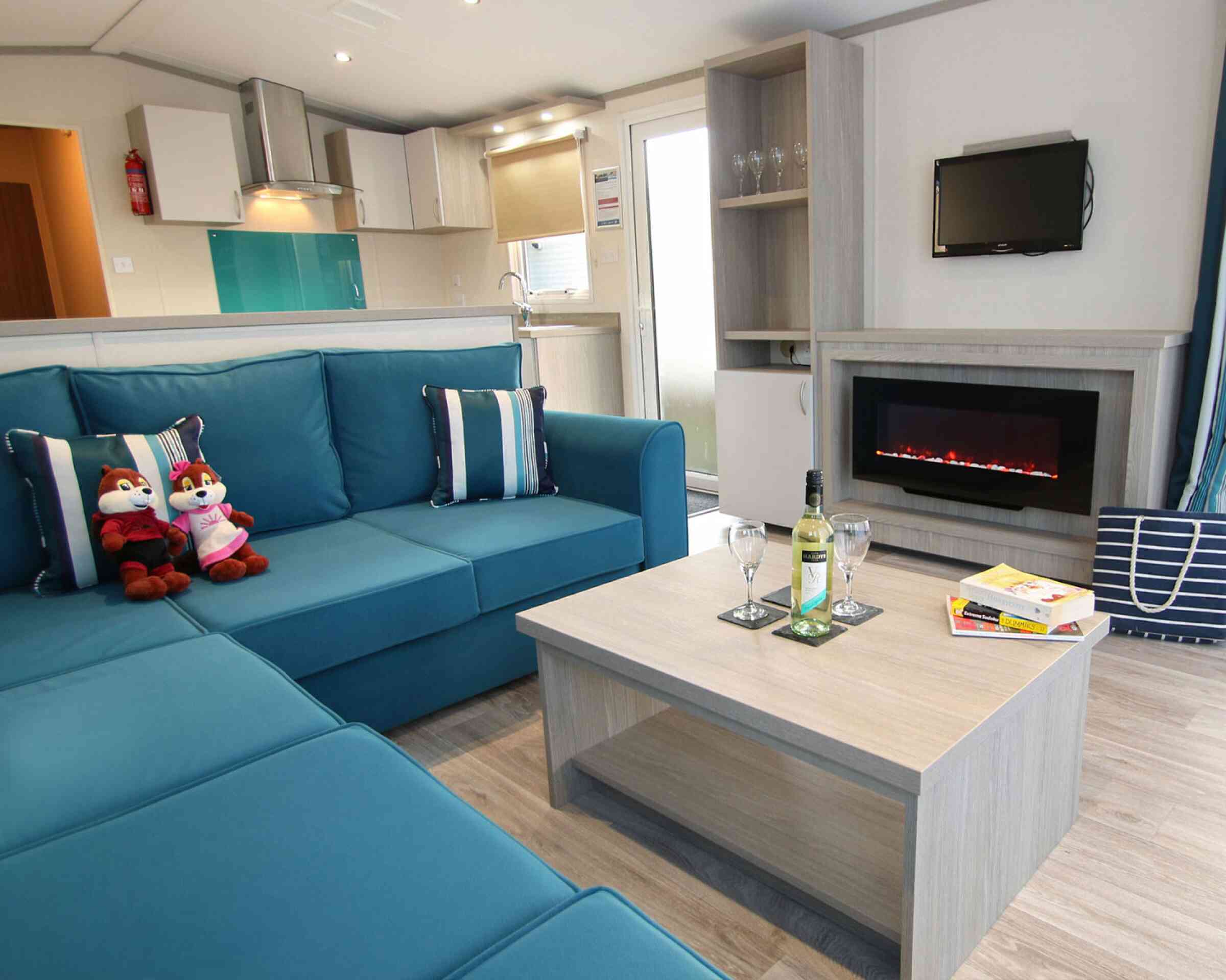 A cozy living room with a teal sectional sofa adorned with blue and white striped cushions. Two plush toys sit on the sofa. A wooden coffee table holds a bottle of wine and two glasses. A modern electric fireplace is below a small wall-mounted TV, with a doorway leading to an outdoor area in the background.