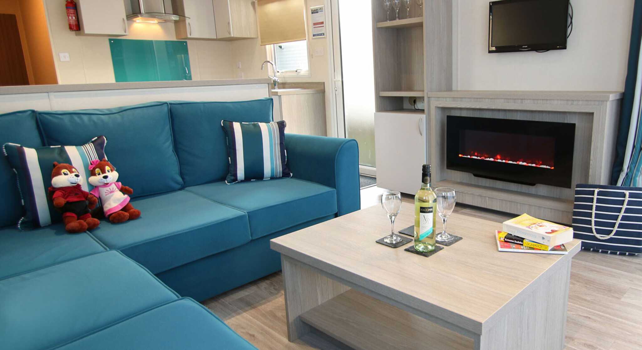 A cozy living room with a teal sectional sofa adorned with blue and white striped cushions. Two plush toys sit on the sofa. A wooden coffee table holds a bottle of wine and two glasses. A modern electric fireplace is below a small wall-mounted TV, with a doorway leading to an outdoor area in the background.