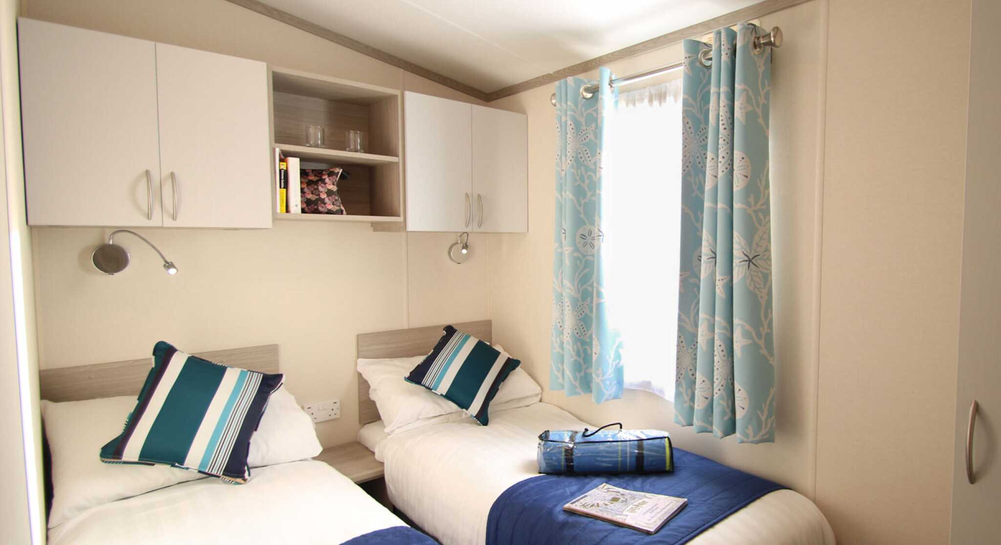 A cozy guest room featuring two single beds with blue and white bedding. There are light-colored walls, a shelf with books, and curtains adorned with a floral pattern. Natural light streams in through a window.