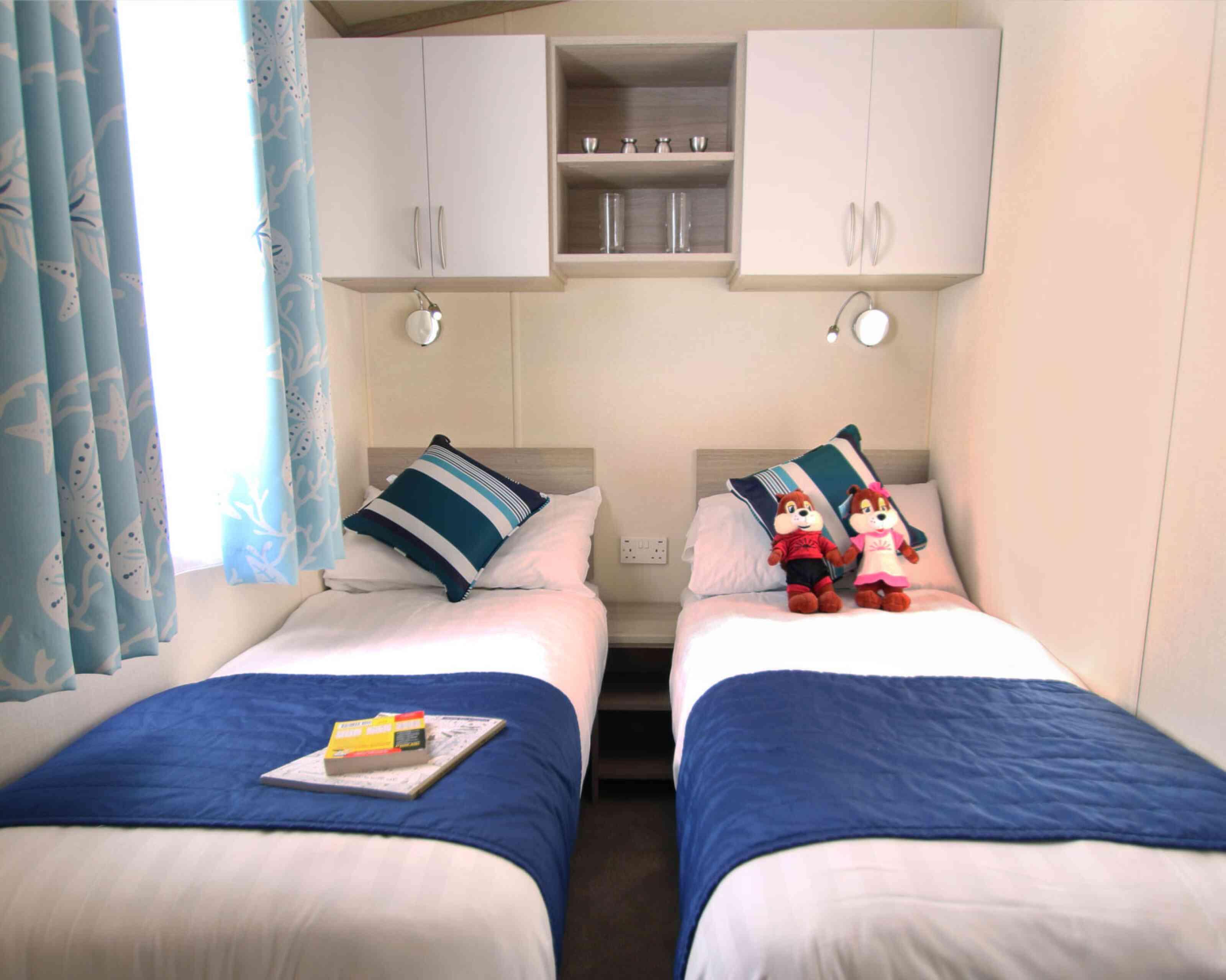 A cozy bedroom featuring two twin beds dressed in blue and white linens. Each bed has matching pillows and a small stuffed animal perched on top. A bedside table holds a magazine and a small bookshelf is mounted above the beds, with a window dressed in light blue curtains.