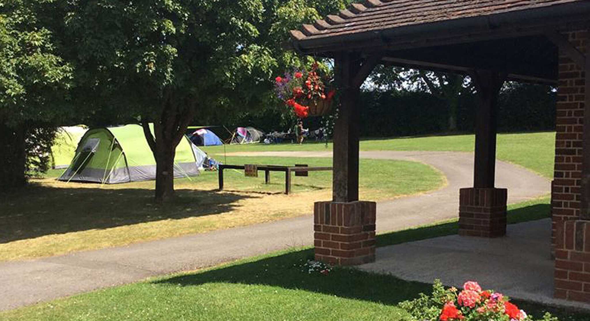 A green camping tent set up on a grassy area, with several other tents in the background. A brick pavilion is in the foreground, featuring hanging flowers and a wooden table. Lush trees surround the campsite, and the sky is clear and blue.