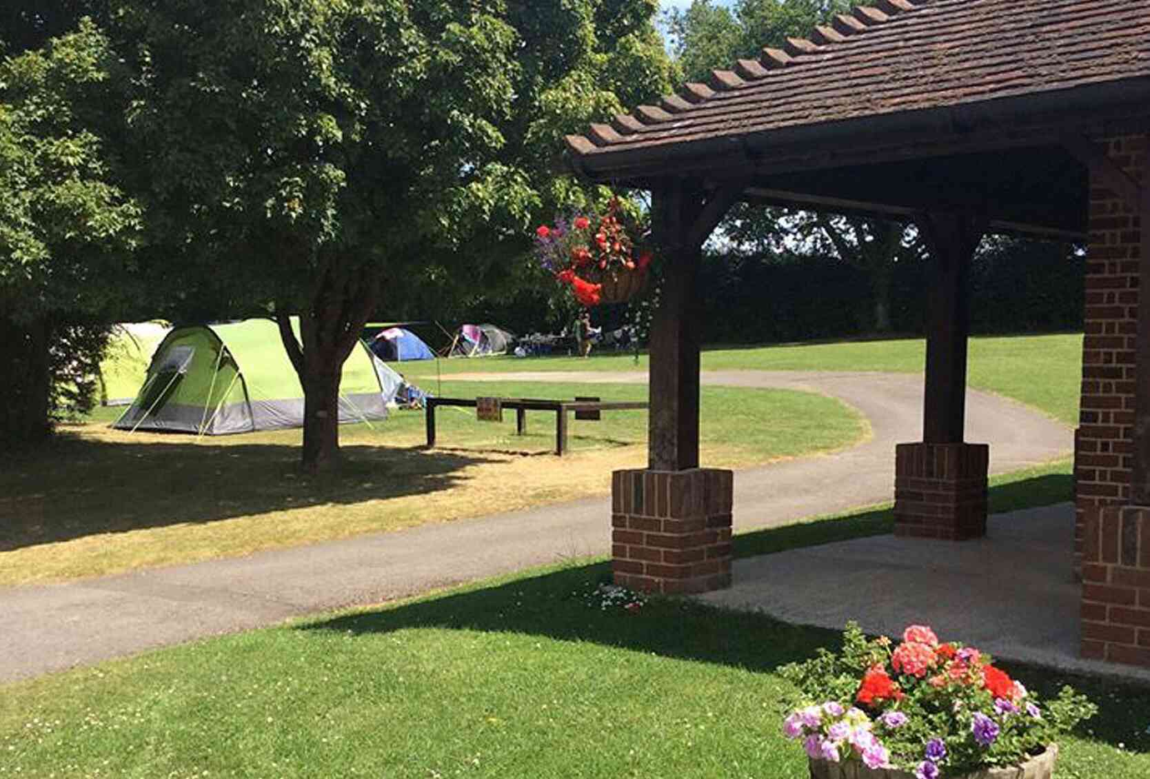 A green camping tent set up on a grassy area, with several other tents in the background. A brick pavilion is in the foreground, featuring hanging flowers and a wooden table. Lush trees surround the campsite, and the sky is clear and blue.