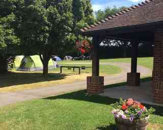 A green camping tent set up on a grassy area, with several other tents in the background. A brick pavilion is in the foreground, featuring hanging flowers and a wooden table. Lush trees surround the campsite, and the sky is clear and blue.