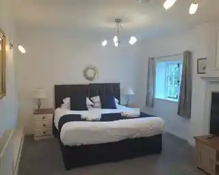A spacious bedroom featuring a large bed with white linens and navy blue accents. There are bedside lamps on either side of the bed, a wooden nightstand, and a decorative mirror on the wall. A window with light curtains provides natural light, and a cozy carpet covers the floor.