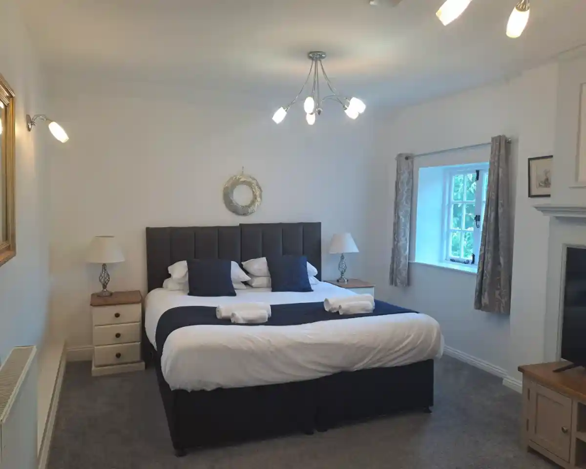 A spacious bedroom featuring a large bed with white linens and navy blue accents. There are bedside lamps on either side of the bed, a wooden nightstand, and a decorative mirror on the wall. A window with light curtains provides natural light, and a cozy carpet covers the floor.