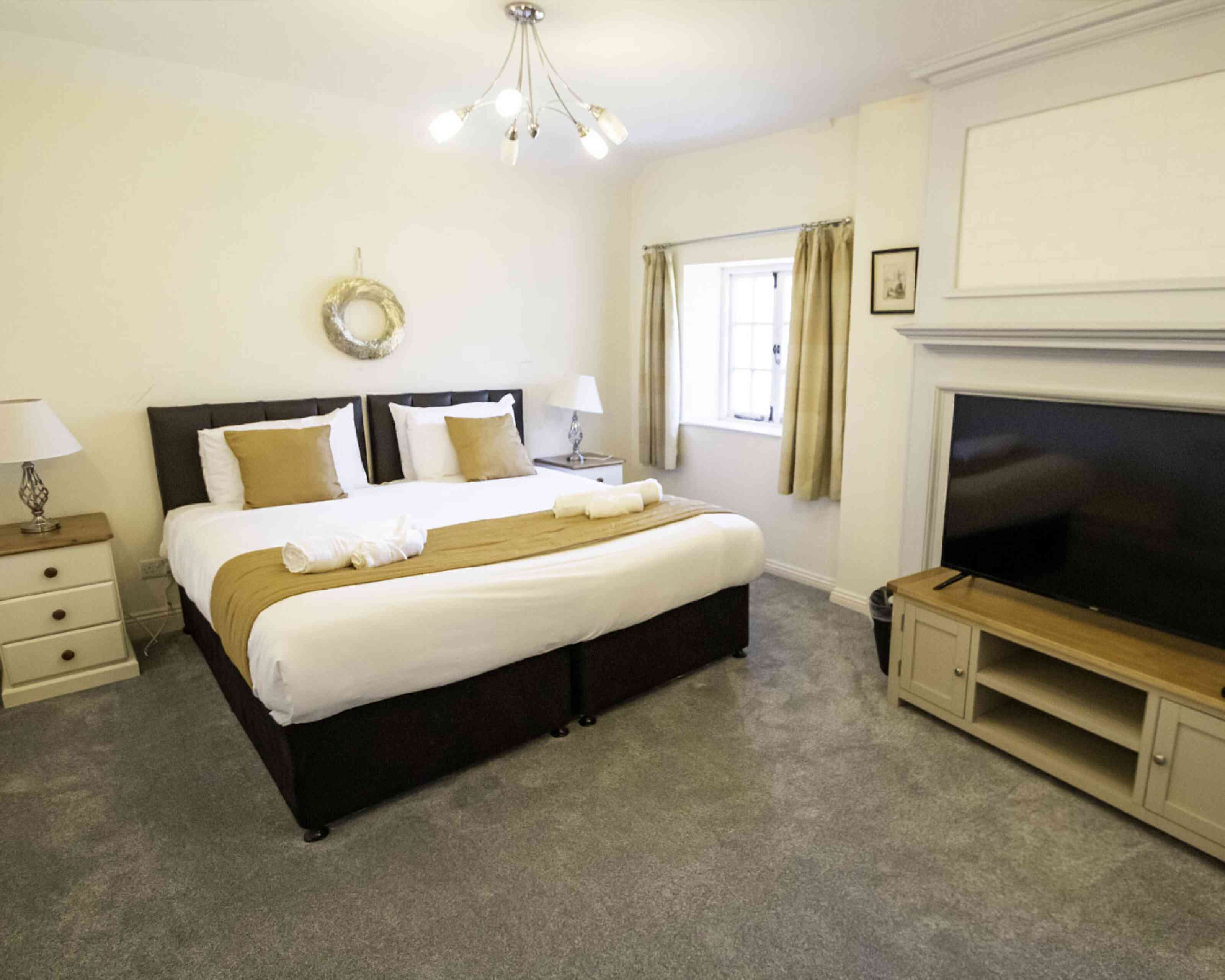 A cozy bedroom featuring a large bed with white and brown bedding, two bedside lamps, and a flat-screen TV mounted on a light wood cabinet. Natural light streams in through a window with drapes.