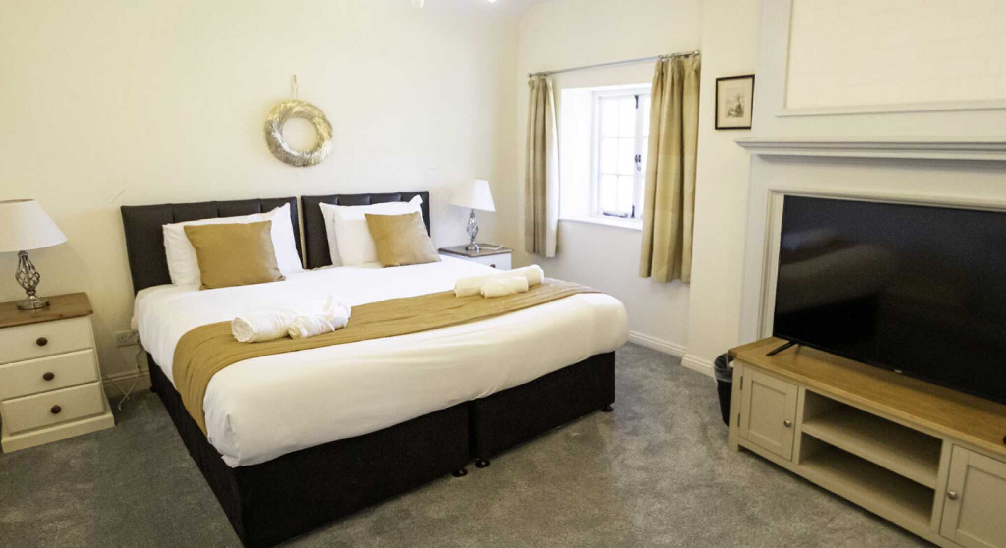 A cozy bedroom featuring a large bed with white and brown bedding, two bedside lamps, and a flat-screen TV mounted on a light wood cabinet. Natural light streams in through a window with drapes.