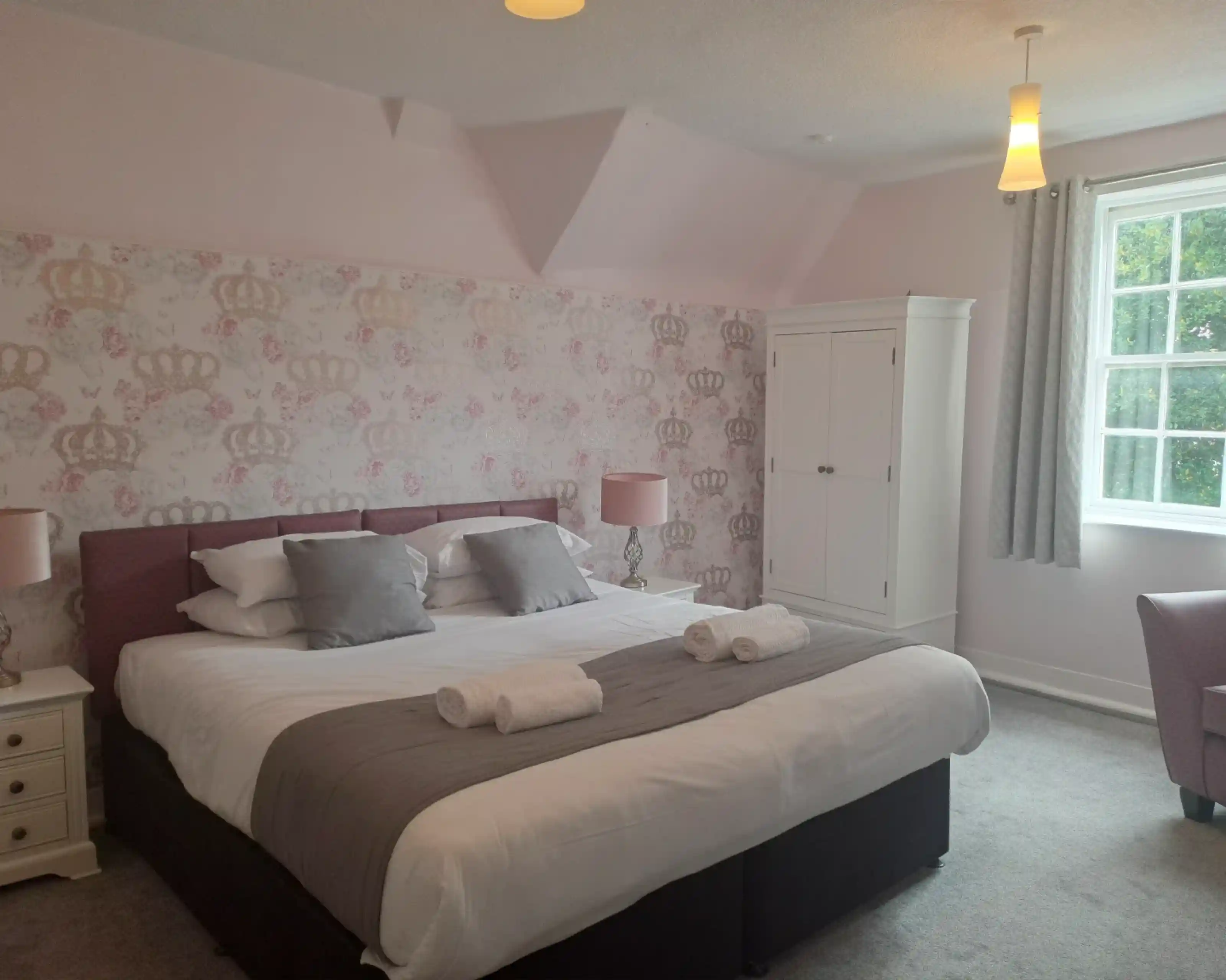 A cozy bedroom featuring a large bed with gray and white bedding, two rolled towels on the bed, and soft pink wallpaper with crown motifs. There is a white wardrobe, a window with curtains, and a cozy chair in the corner, creating a welcoming atmosphere.