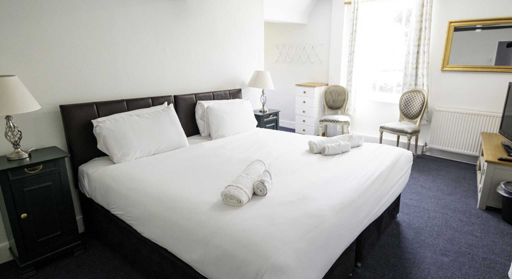 A spacious bedroom featuring a large bed with white linens, flanked by two bedside tables with lamps. A mirror hangs on the wall opposite a window with sheer curtains, and a dresser sits nearby. Two elegant chairs add a touch of decor.