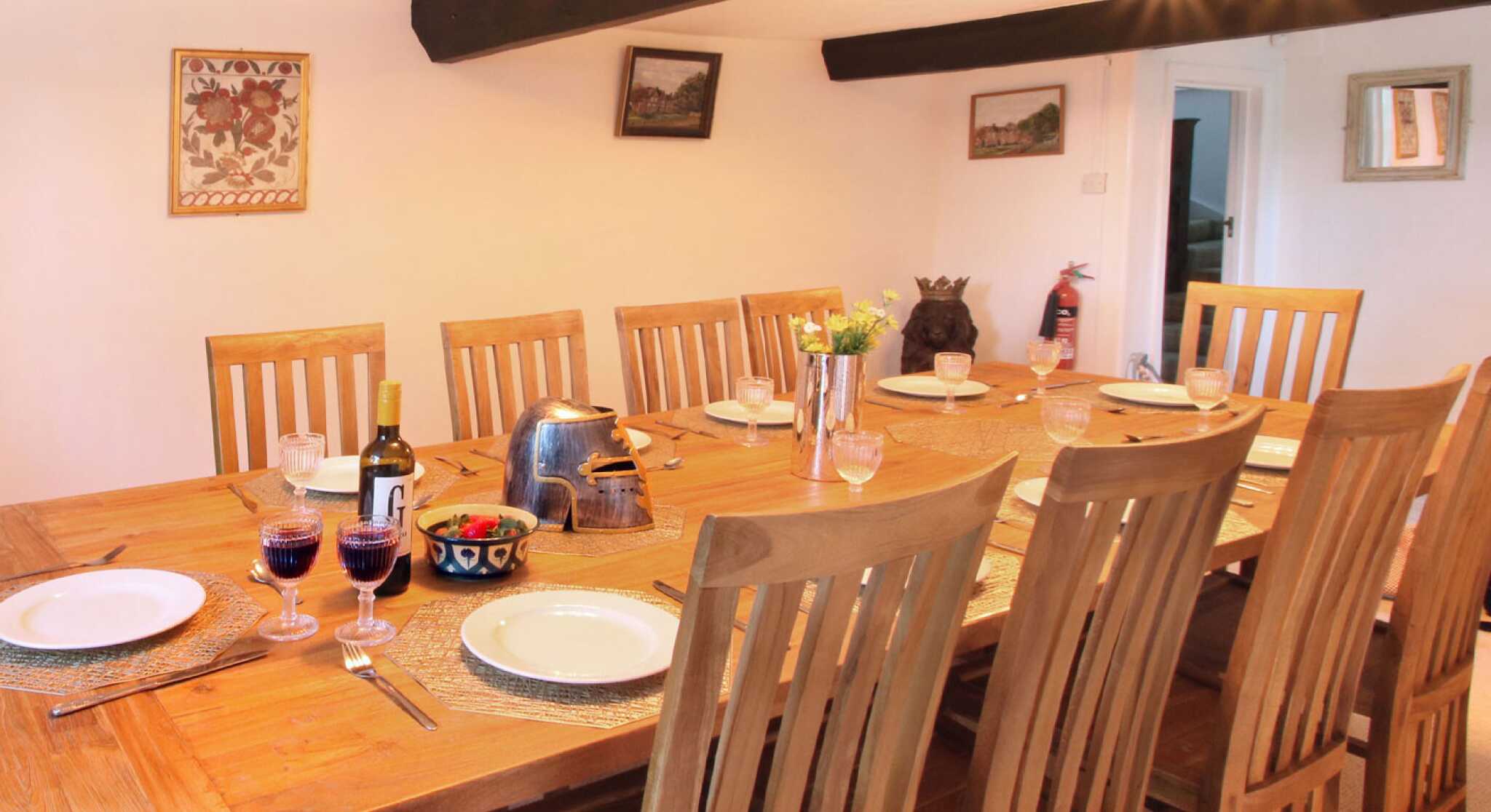 A rustic dining room featuring a long wooden table set with plates and glasses. Nearby, a bottle of wine and glasses filled with red liquid are visible. The space has wooden beams on the ceiling and decorative wall art.