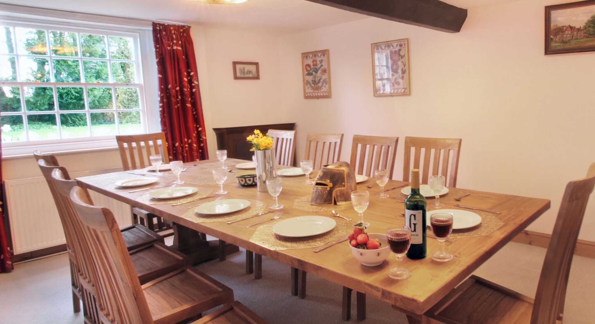 A spacious dining room featuring a large wooden table set for a meal, with plates, glassware, and decorative items. The room is brightened by windows and has warm lighting, complemented by red curtains and framed artwork on the walls.
