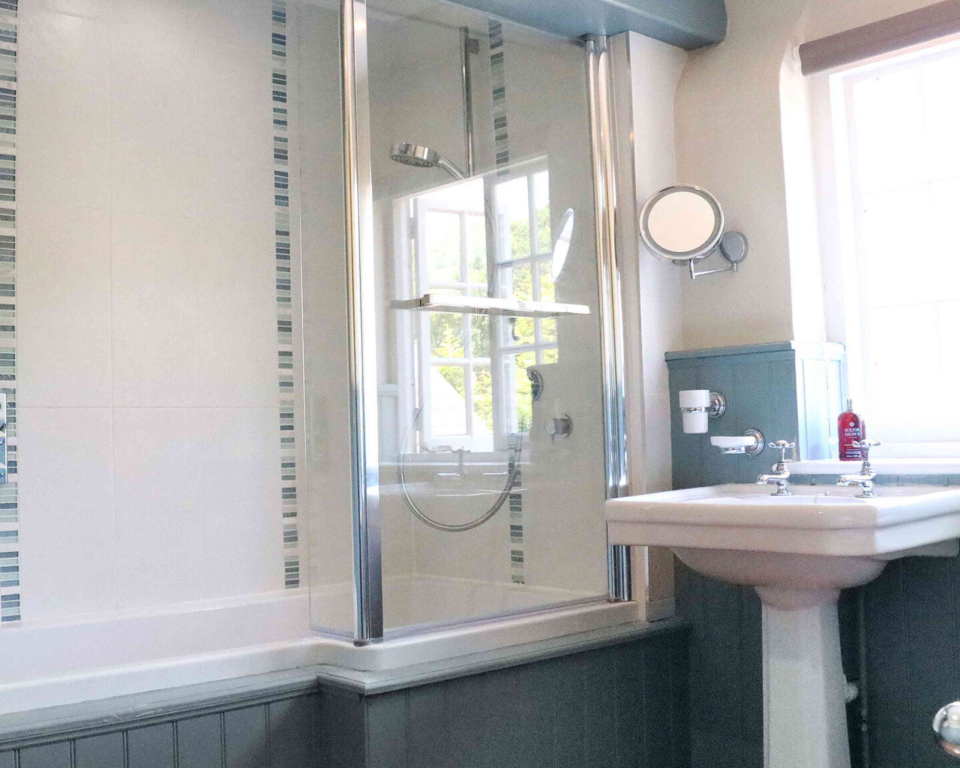 A modern bathroom featuring a glass shower enclosure, a bathtub, and a sink with traditional fittings. The walls are partially tiled with decorative strips, and a window allows natural light to enter.