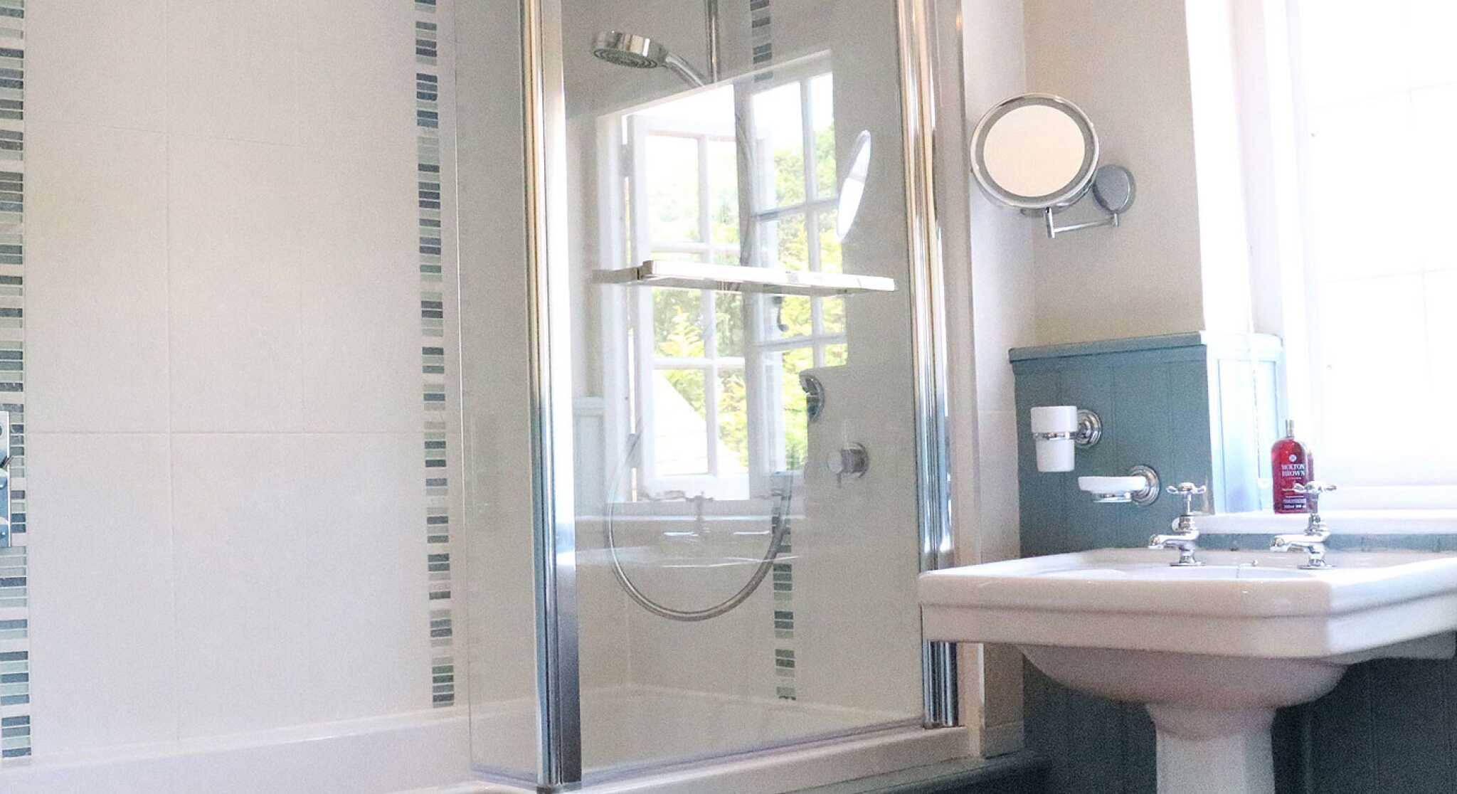 A modern bathroom featuring a glass shower enclosure, a bathtub, and a sink with traditional fittings. The walls are partially tiled with decorative strips, and a window allows natural light to enter.